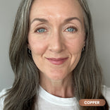 A woman with long, gray-streaked hair and blue eyes smiles gently at the camera. She wears a white shirt and natural makeup featuring PrimePrometics™ PrimeEyes Glide *New* Bundle smudge-proof eyeliner in “COPPER.”.