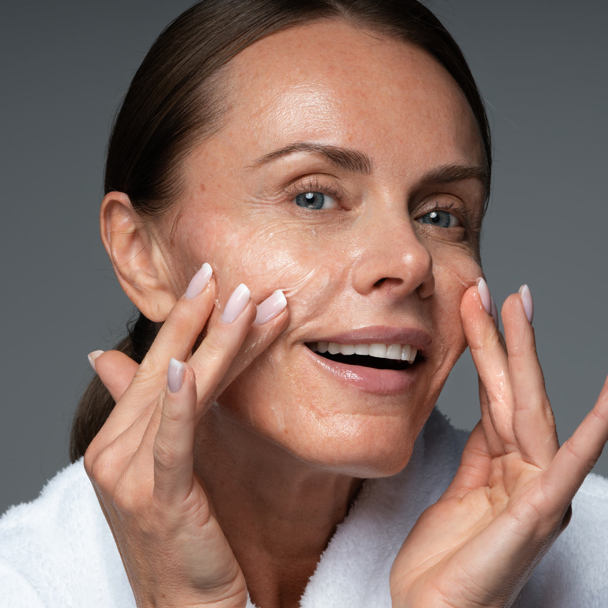 Mature woman in a white robe massaging Prime Prometics Gentle Gel Cleanser foam onto her cheeks