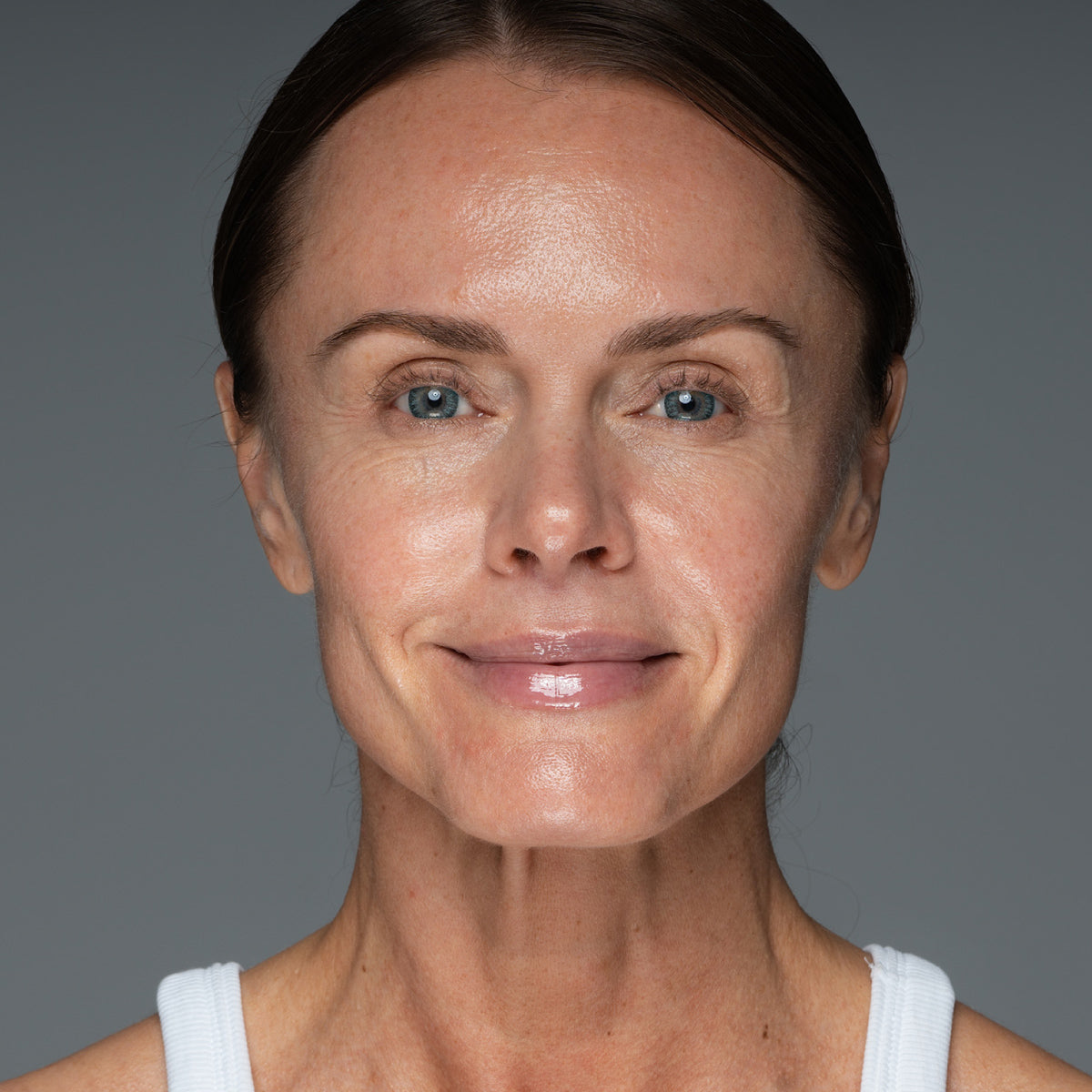 A middle-aged woman with light skin, blue eyes, and brown hair pulled back smiles gently at the camera. She is wearing a white tank top and is set against a plain gray background.