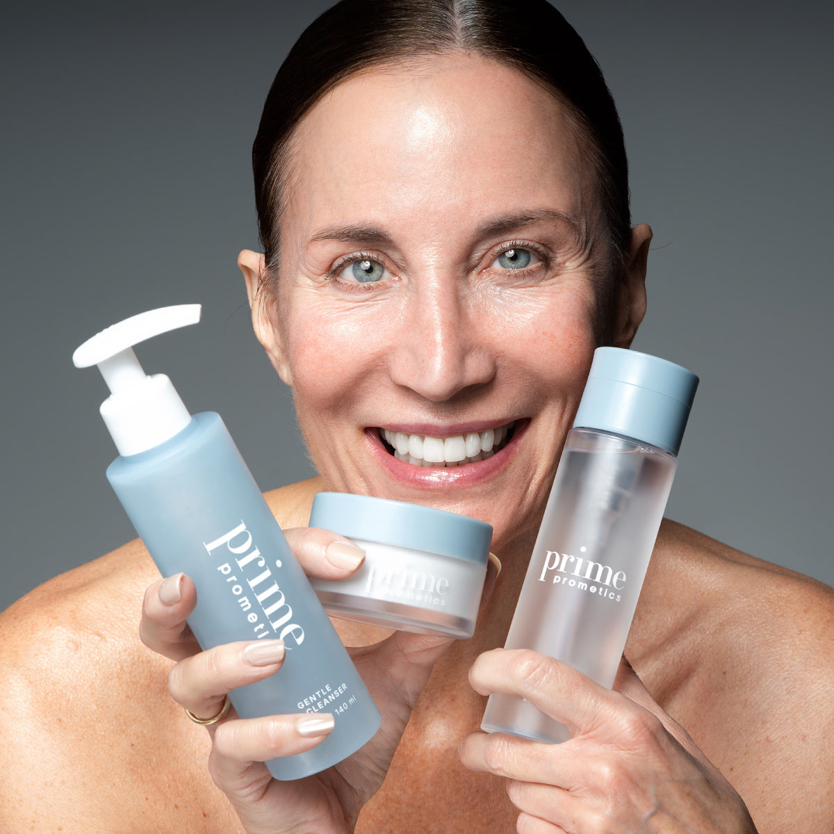 A smiling woman holds up three blue and white skincare products labeled prime prometic against a gray background. She appears happy and confident, showcasing the cleanser, cream, and toner.
