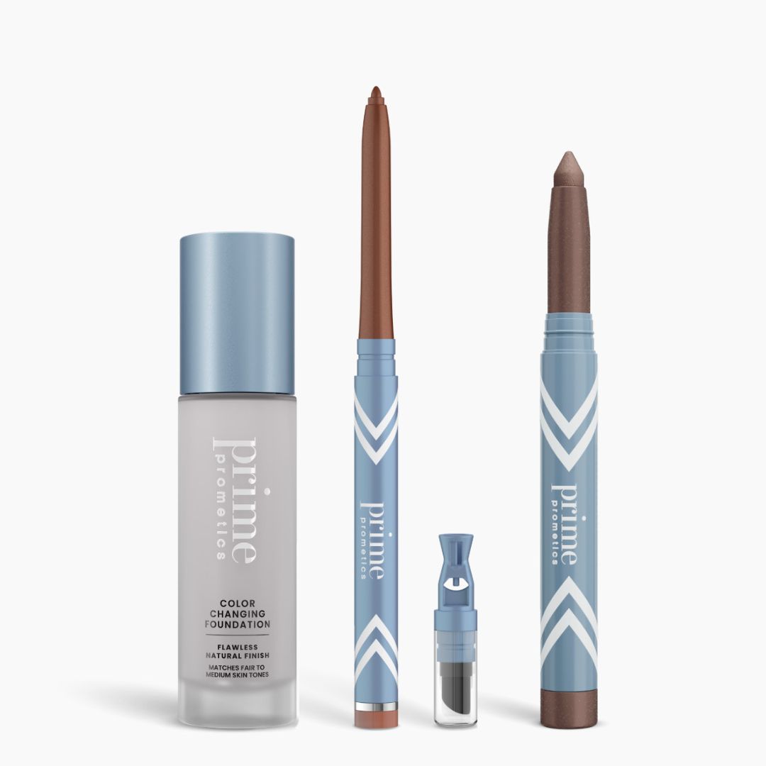 Four blue and silver PrimePrometics™ All-Day Confidence Bundle products stand upright: a color-changing foundation bottle, brown glide eyeliner pencil, small applicator vial, and thick brown eyeshadow stick.