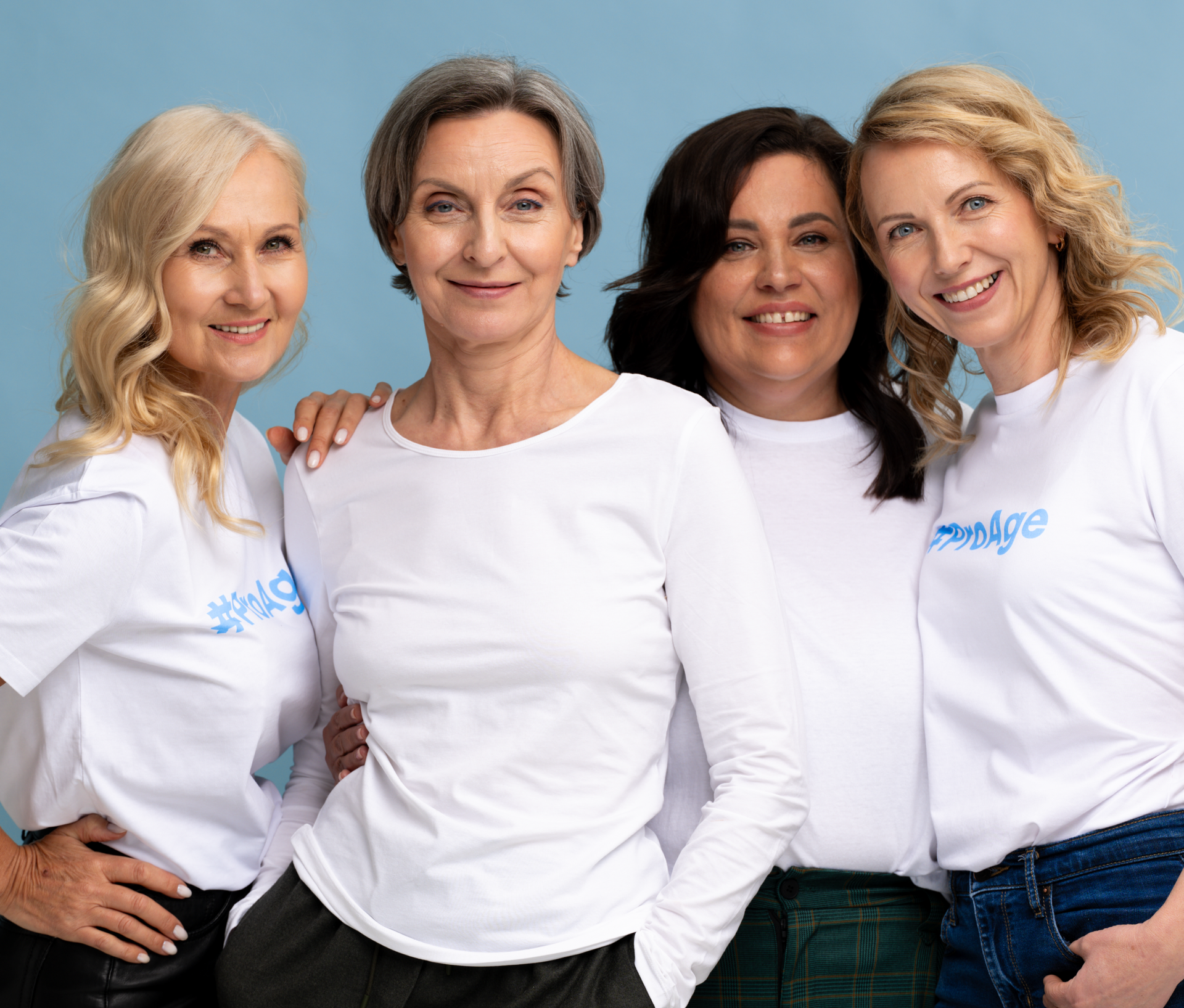 Four smiling women with light hair, dressed in white long-sleeve shirts, stand close together against a blue background, showing a sense of friendship and support.