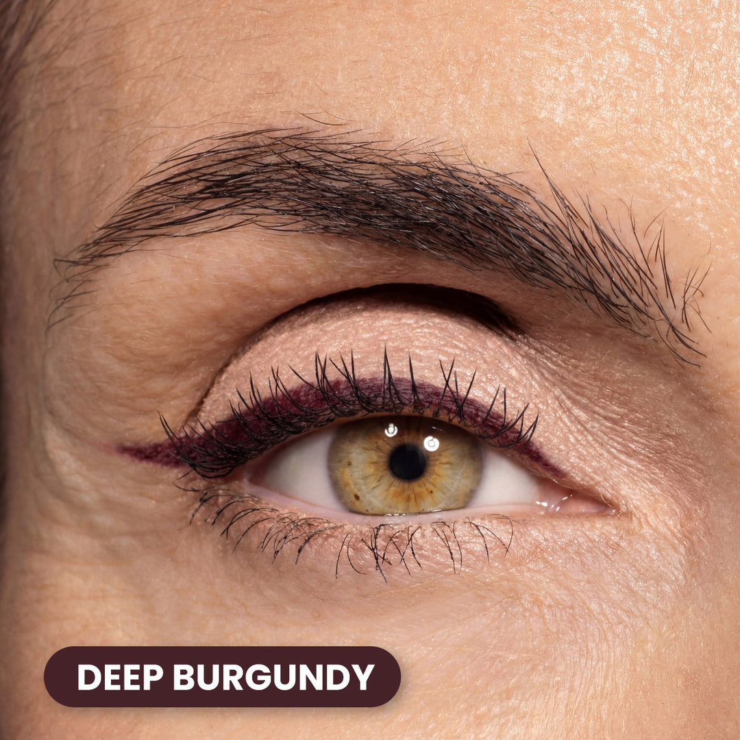 Close-up of a persons eye with hazel iris, burgundy eyeliner along the upper lash line, and well-groomed eyebrow. A label in the bottom left corner reads DEEP BURGUNDY in white text on a burgundy background.