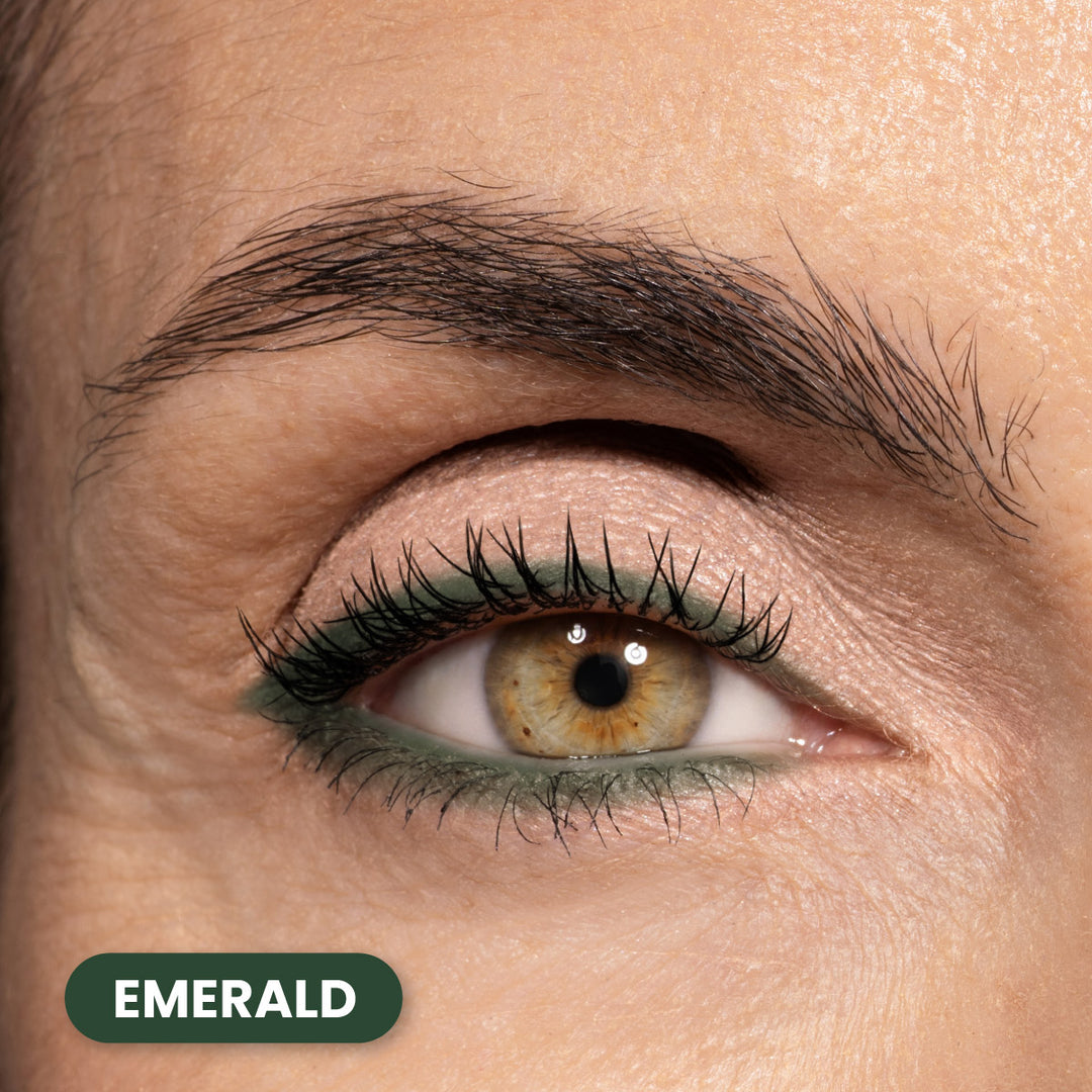 A close-up of a persons eye with green eyeliner on the lower lash line. The eye is hazel, and the skin is fair. There is a label in the bottom left corner that says EMERALD in white text on a green background.