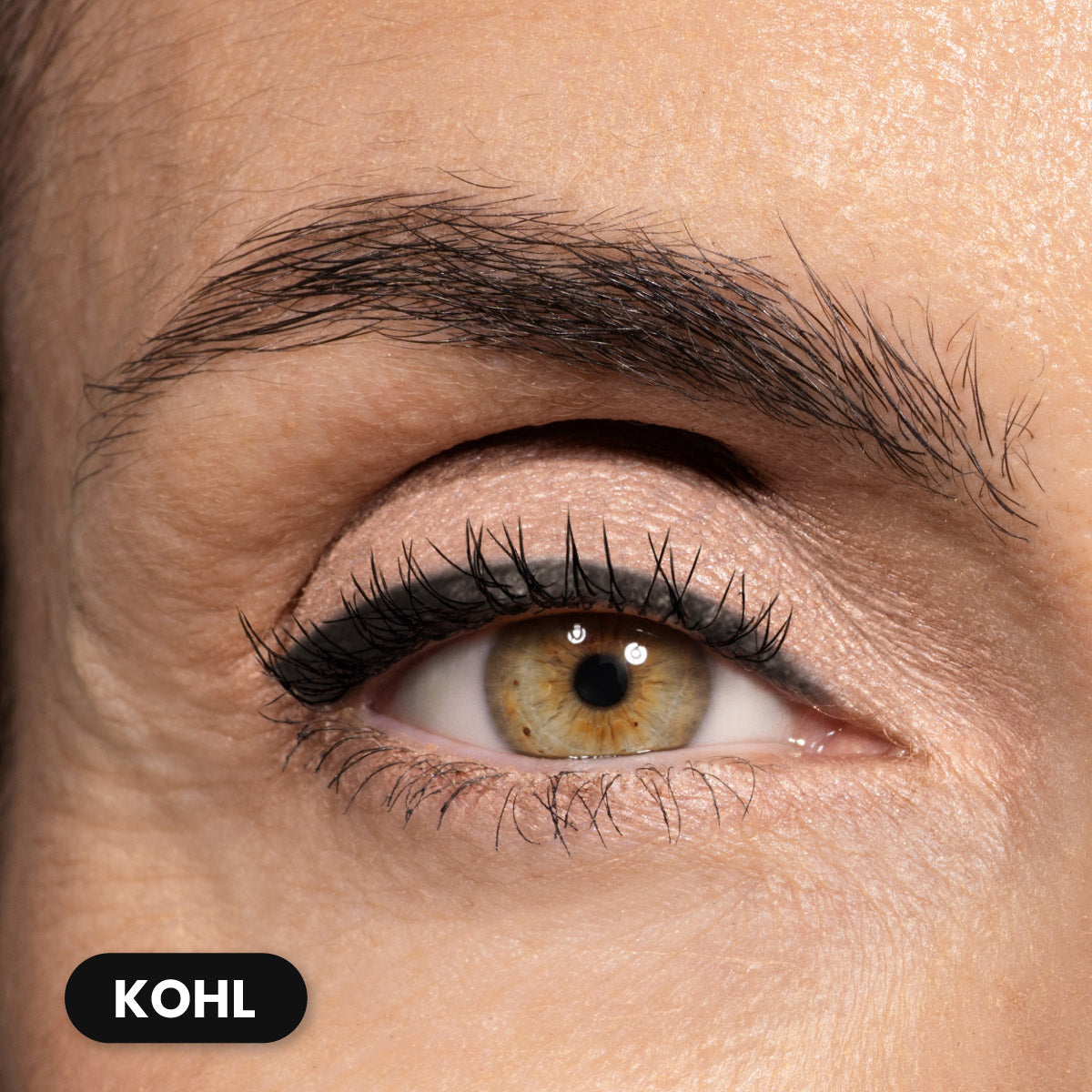 A close-up of a person’s eye with light brown iris, defined eyebrow, and black kohl eyeliner along the upper lash line. The skin is fair and the lashes are long and separated. A label in the corner reads KOHL.