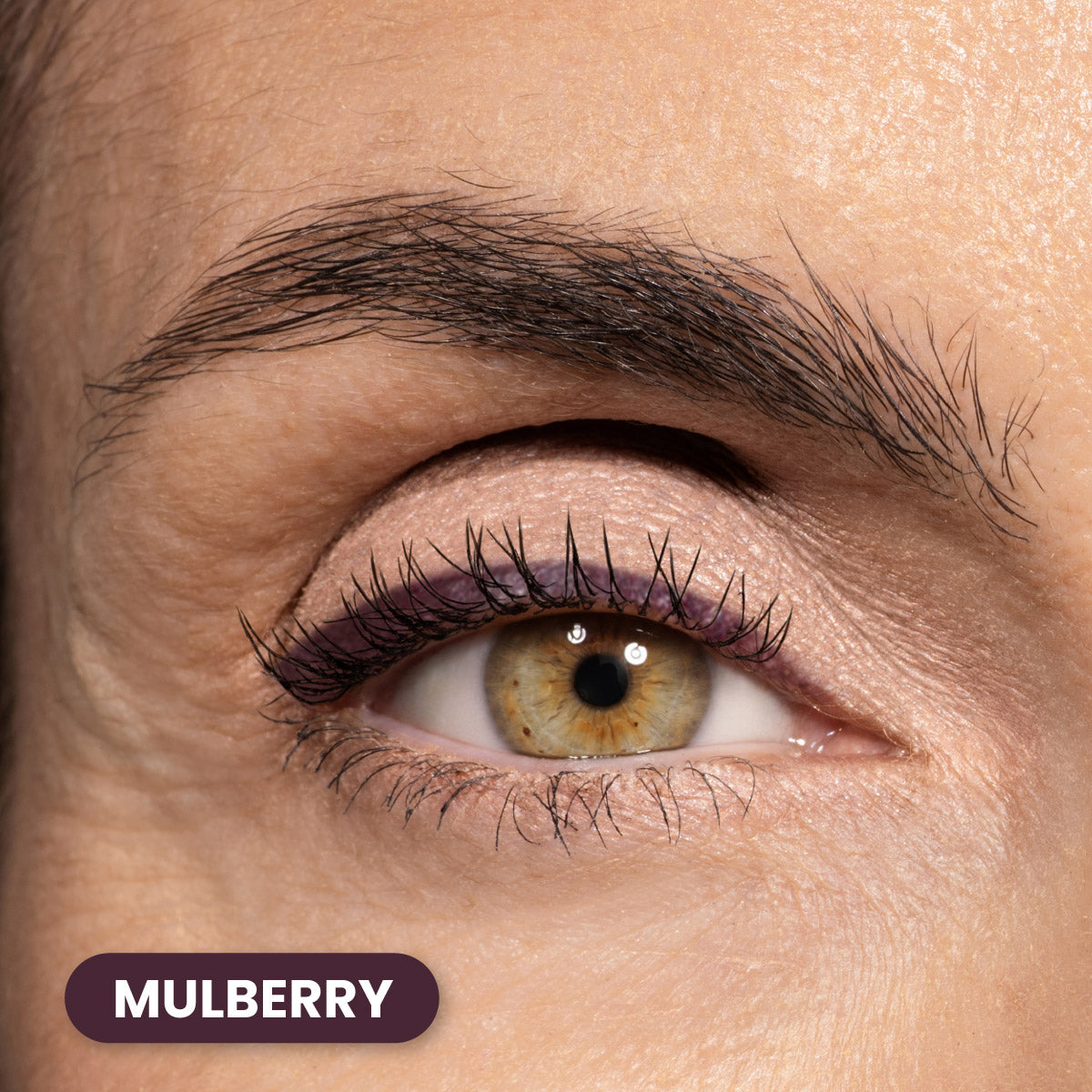 A close-up of a persons eye with light brown hazel iris, subtle purple eyeliner, and well-groomed eyebrow. The word MULBERRY appears in a dark purple rounded rectangle at the bottom left.