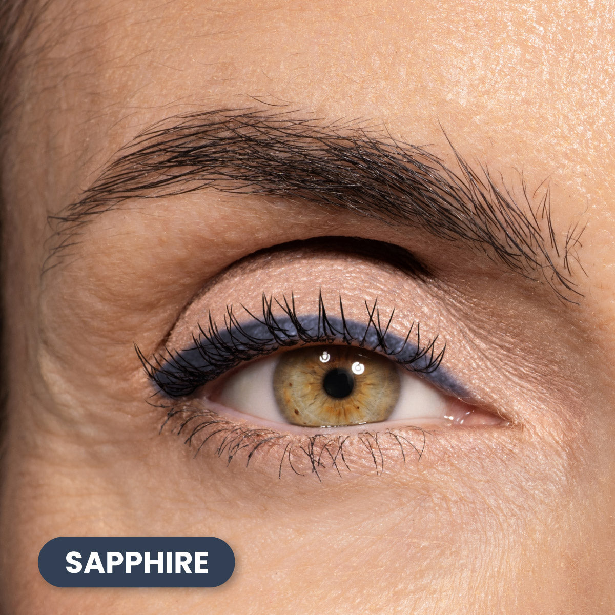 Close-up of a persons eye with brown iris, natural eyelashes, and blue eyeliner on the upper eyelid. The word SAPPHIRE appears in a blue rounded rectangle at the bottom left of the image.