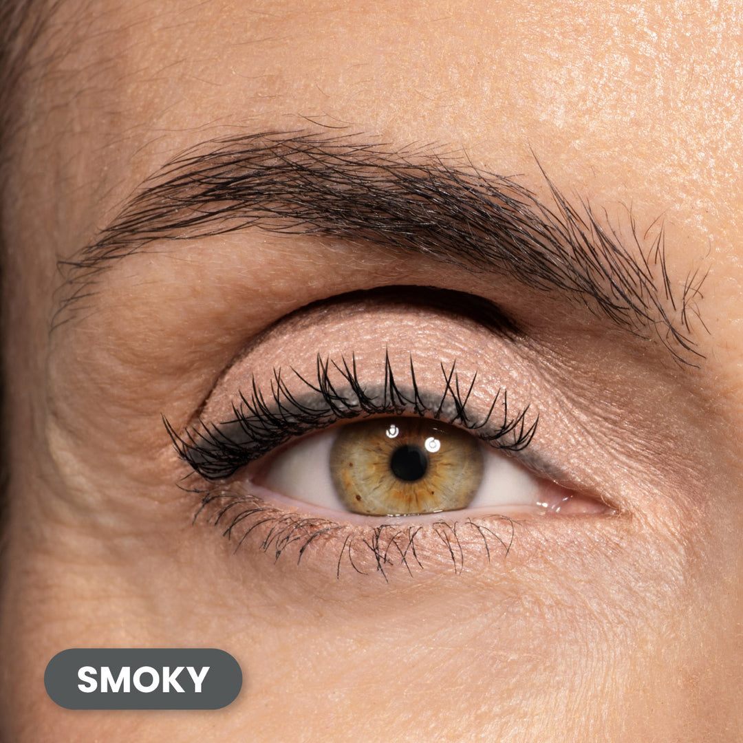 A close-up of a person’s brown eye with defined lashes and natural nude eyeshadow. The eyebrow is full and neatly groomed. A label in the bottom left corner reads SMOKY.