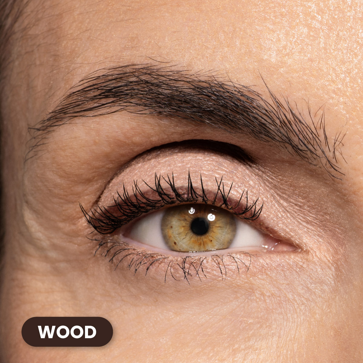 A close-up of a light brown eye with long lashes and natural makeup. The skin is fair with visible texture, and the word WOOD appears in a rounded dark label in the lower left corner.