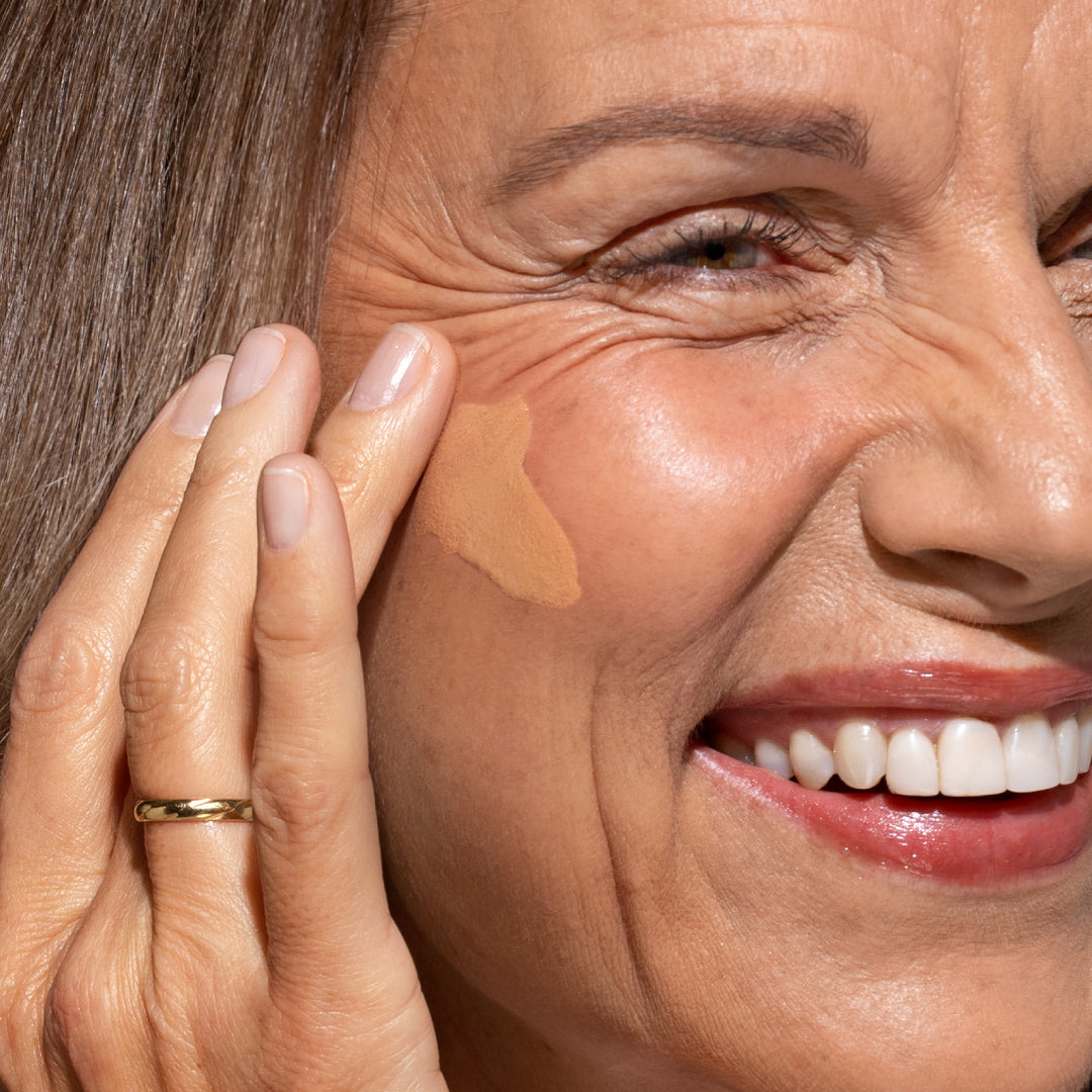 A close-up of a smiling woman applying liquid foundation to her cheek with her fingers. She has light skin, visible wrinkles, and is wearing a gold ring on her finger.
