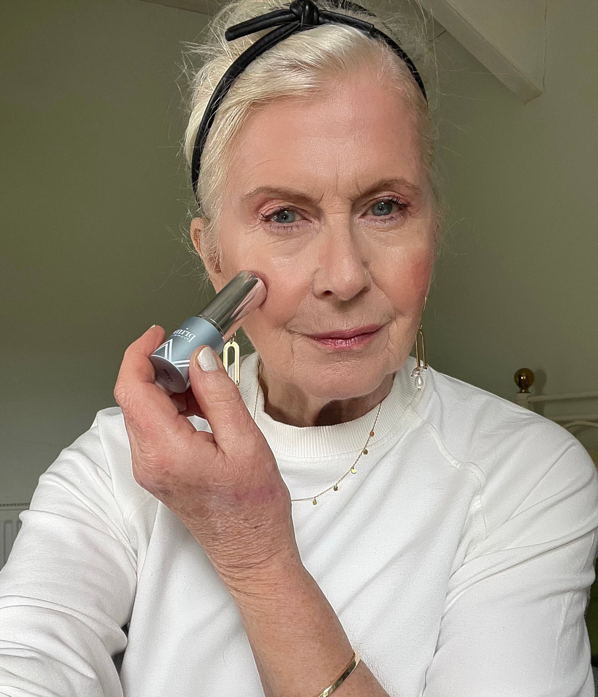 An older woman with light hair, tied back with a black headband, applies makeup to her cheek using a stick applicator. She is wearing a white top, gold jewelry, and is looking at the camera.
