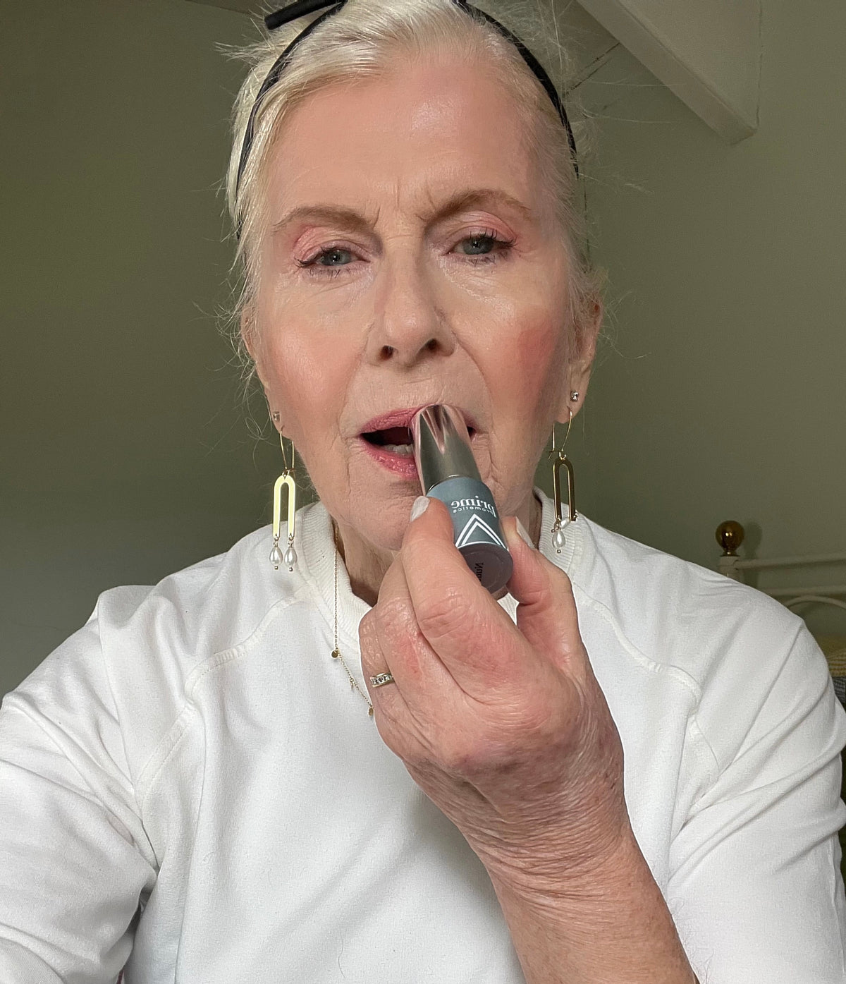 An older woman with light skin and white hair applies pink lipstick while wearing a white top, gold earrings, and a black hairband. She is indoors, facing the camera, in natural light.