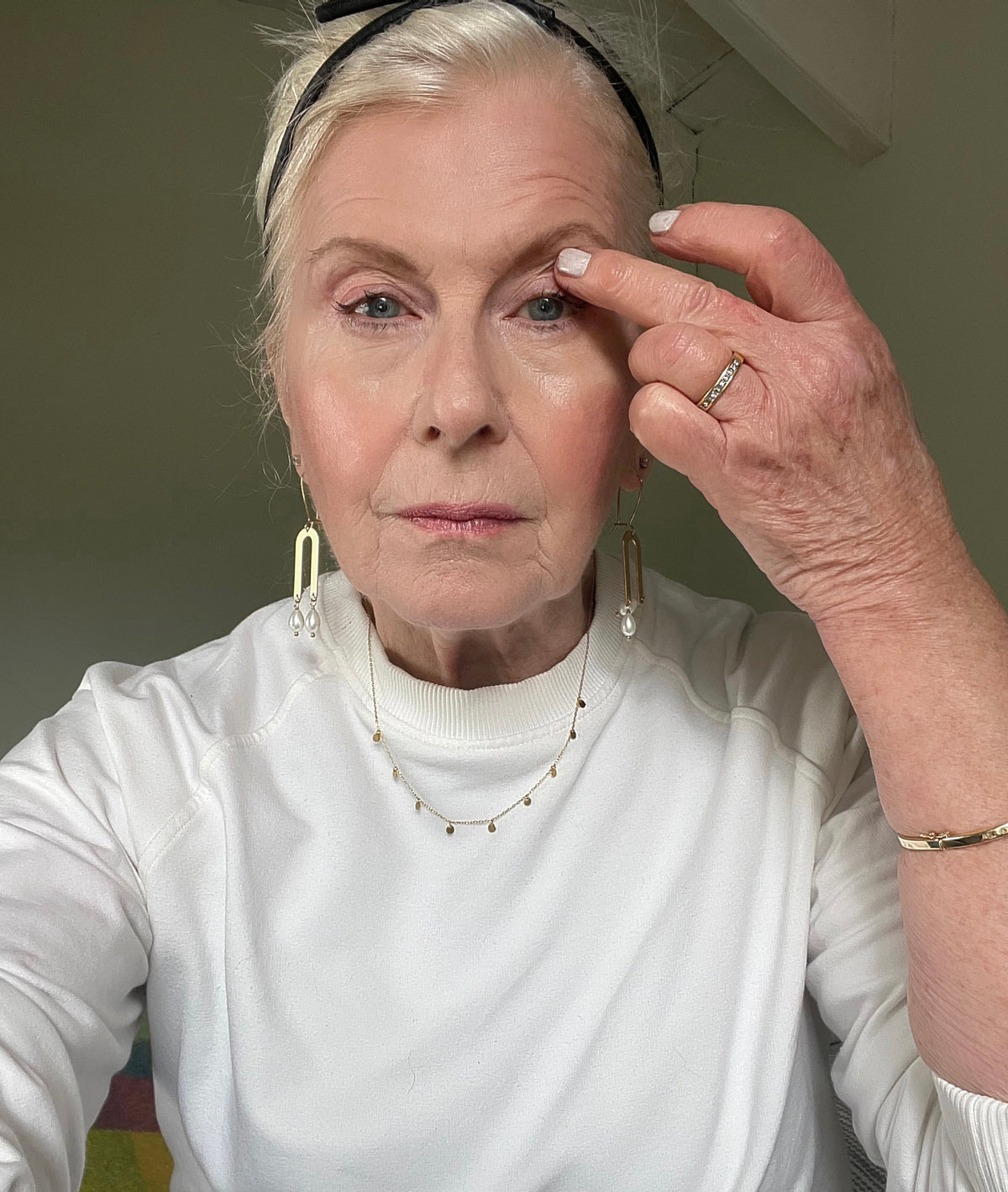 An older woman with light hair and fair skin, wearing a white top and gold jewelry, touches her right eyelid with her index finger while looking directly at the camera.