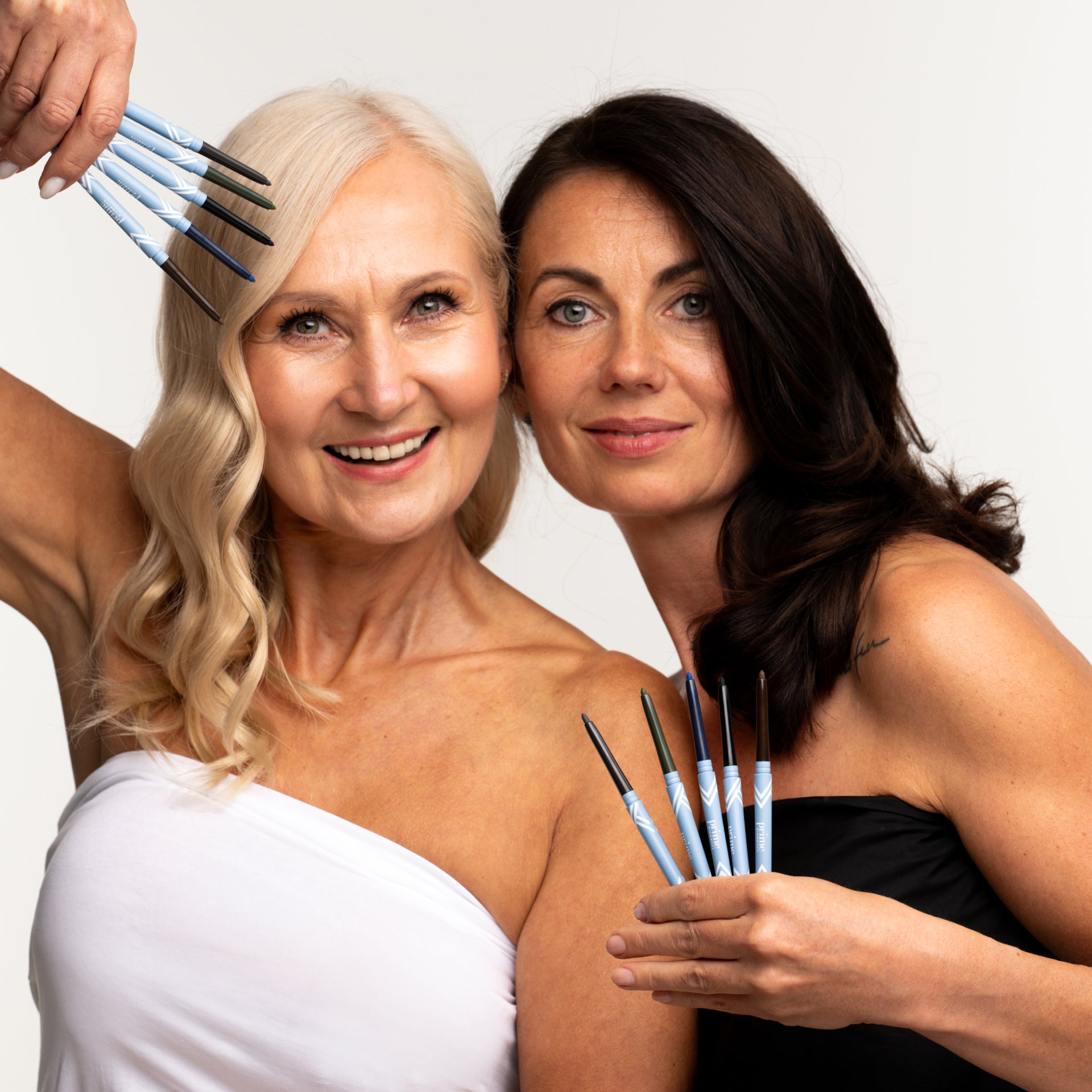 Two women with light skin, one with blonde hair and one with dark hair, smile at the camera while holding blue makeup brushes near their faces. They wear sleeveless tops and pose against a plain white background.