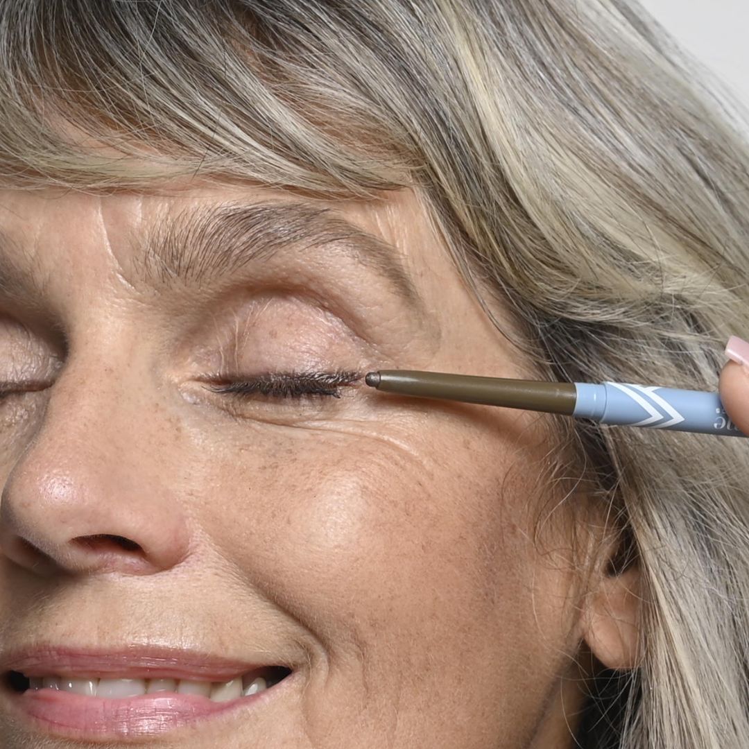 A close-up of an older woman smiling with her eyes closed as she applies brown eyeliner to her upper eyelid. Her skin and hair are light, and she appears happy and relaxed.
