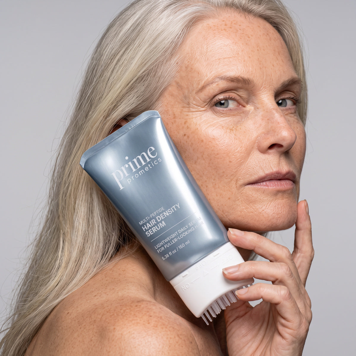 Eine Frau mit langen grauen Haaren hält eine Tube „PrimePrometics™ Multi-Peptide Hair Density Serum“ mit integriertem Applikatorpinsel nahe an ihr Gesicht und präsentiert das Produkt, das für volleres Haar sorgen soll.