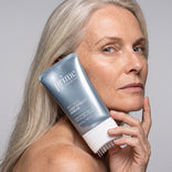 Eine Frau mit langen grauen Haaren hält eine Tube „PrimePrometics™ Multi-Peptide Hair Density Serum“ mit integriertem Applikatorpinsel nahe an ihr Gesicht und präsentiert das Produkt, das für volleres Haar sorgen soll.