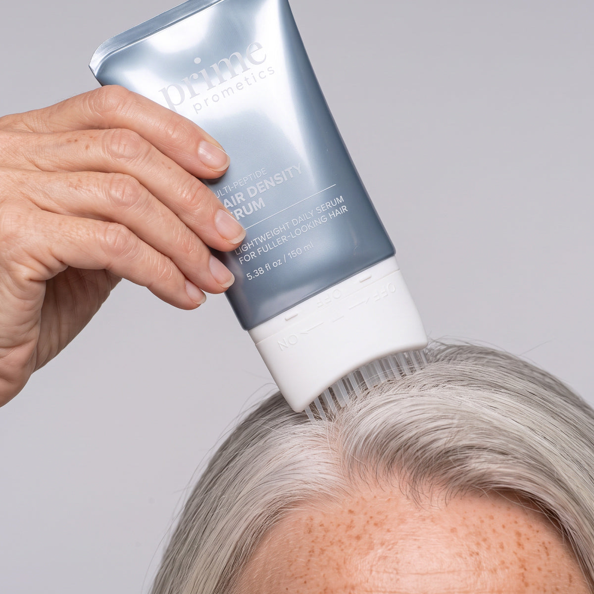 Eine Hand trägt das PrimePrometics™ Multi-Peptide Hair Density Serum aus einer silbernen Tube mit Pinselspitze auf die Kopfhaut einer Person mit grauen Haaren auf, um für volleres Haar zu sorgen.