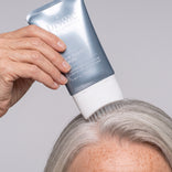 Eine Hand trägt das PrimePrometics™ Multi-Peptide Hair Density Serum aus einer silbernen Tube mit Pinselspitze auf die Kopfhaut einer Person mit grauen Haaren auf, um für volleres Haar zu sorgen.