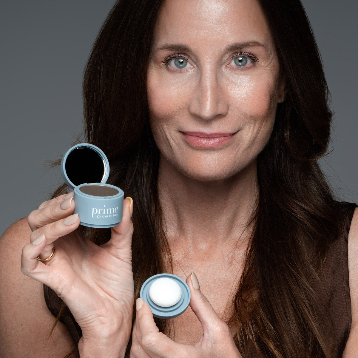 A woman with long brown hair smiles and holds up a small blue cosmetic container with a mirror and a white applicator sponge, labeled prime.