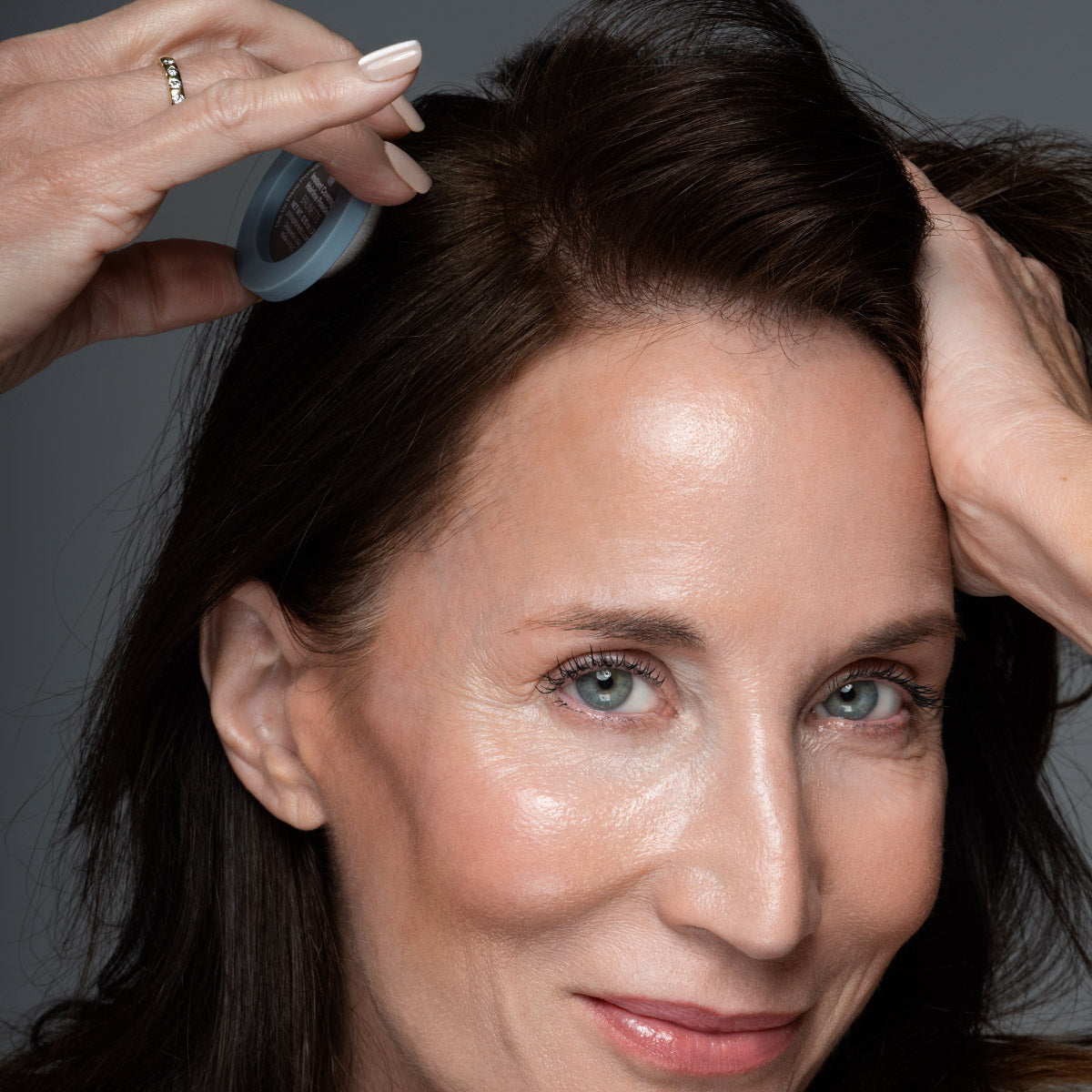 A smiling woman with fair skin uses a round applicator to apply product to her brown hair near the roots, with one hand lifting her hair.