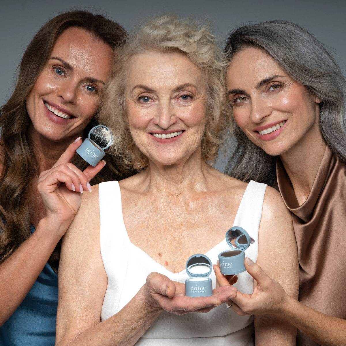 Three smiling women of different ages pose together, each holding a small container labeled prime. They stand close, showcasing the product and exuding confidence and happiness against a simple gray background.