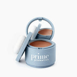 A light blue round container labeled PrimePrometics™ holds Instant Coverage Hairline Powder, featuring a pressed formula, an attached mirror, and a white applicator puff resting on the lid.