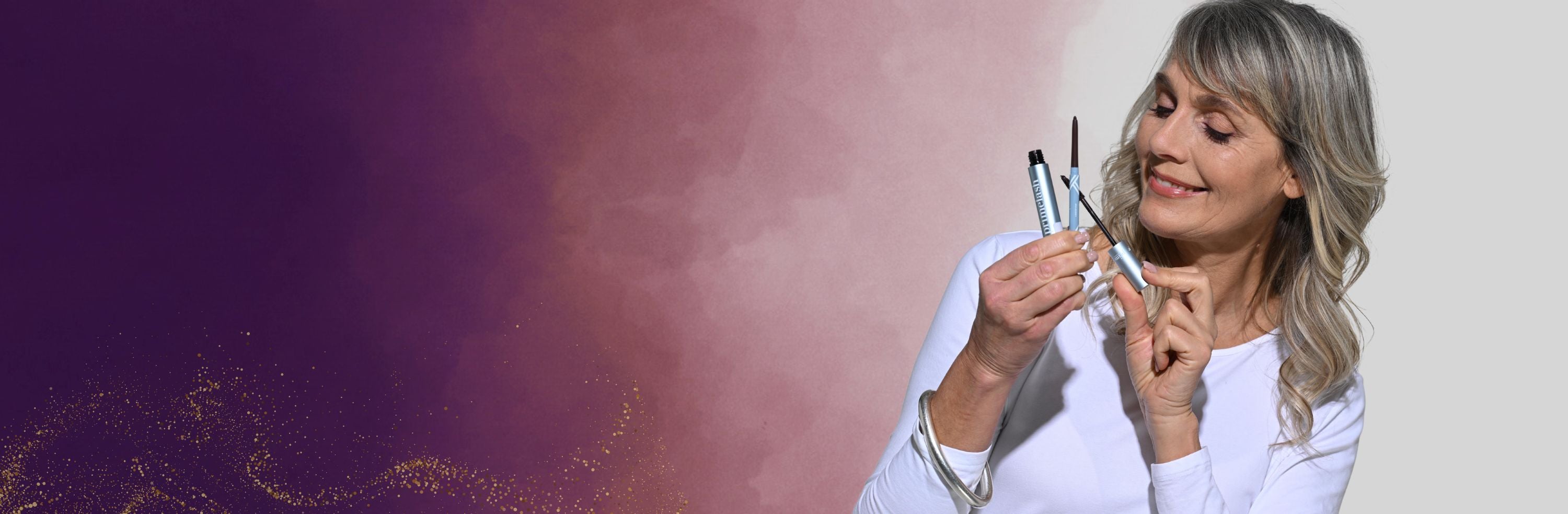 A smiling woman with gray hair wearing a white top holds and examines makeup products against a background blending purple, pink, and beige tones.