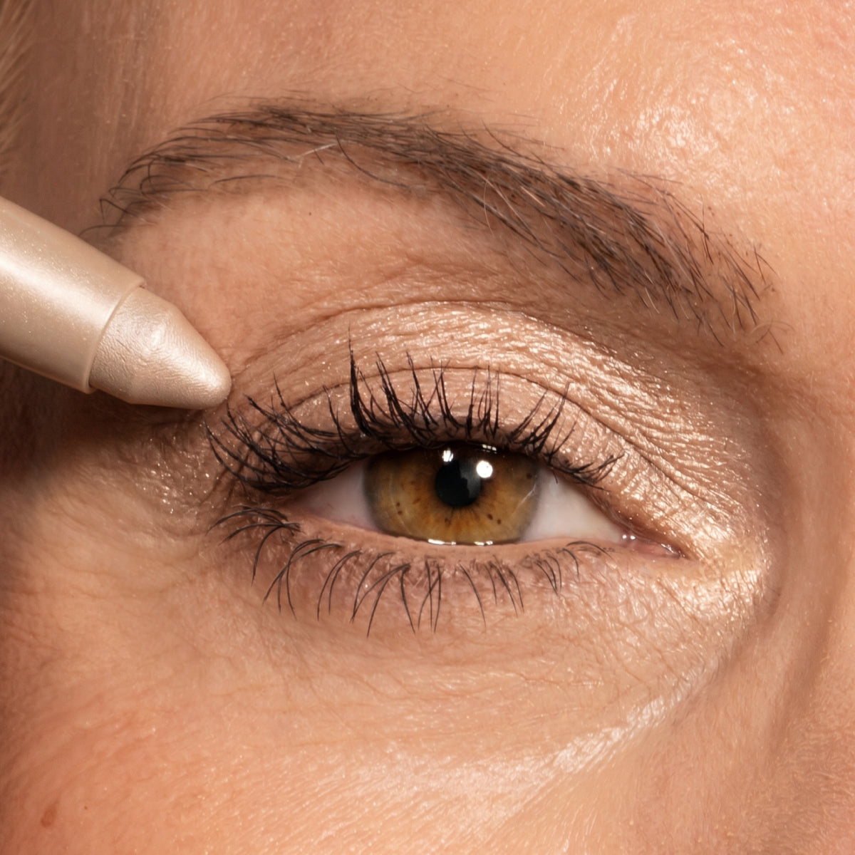 Eine Nahaufnahme eines Auges mit braunem und goldenem Lidschatten. Am äußeren Augenwinkel des oberen Augenlids wird ein Augenstift aufgetragen. Details der Augenbrauen und der Haut sind zu erkennen.