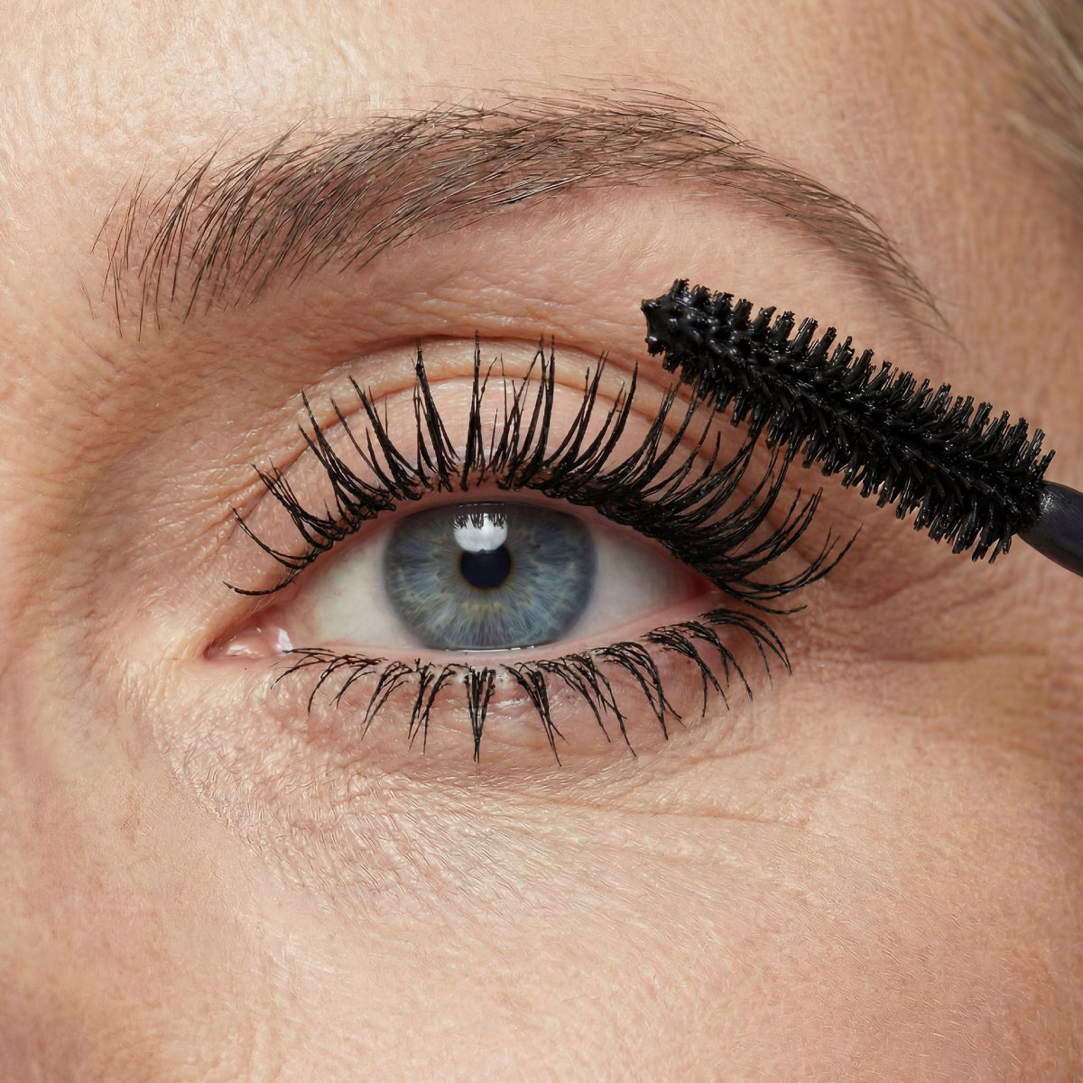 Eine Nahaufnahme eines blauen Auges, bei der mit einem schwarzen Mascara-Bürstchen Mascara auf die oberen Wimpern aufgetragen wird. Die Haut um das Auge herum weist eine natürliche Struktur und feine Fältchen auf.