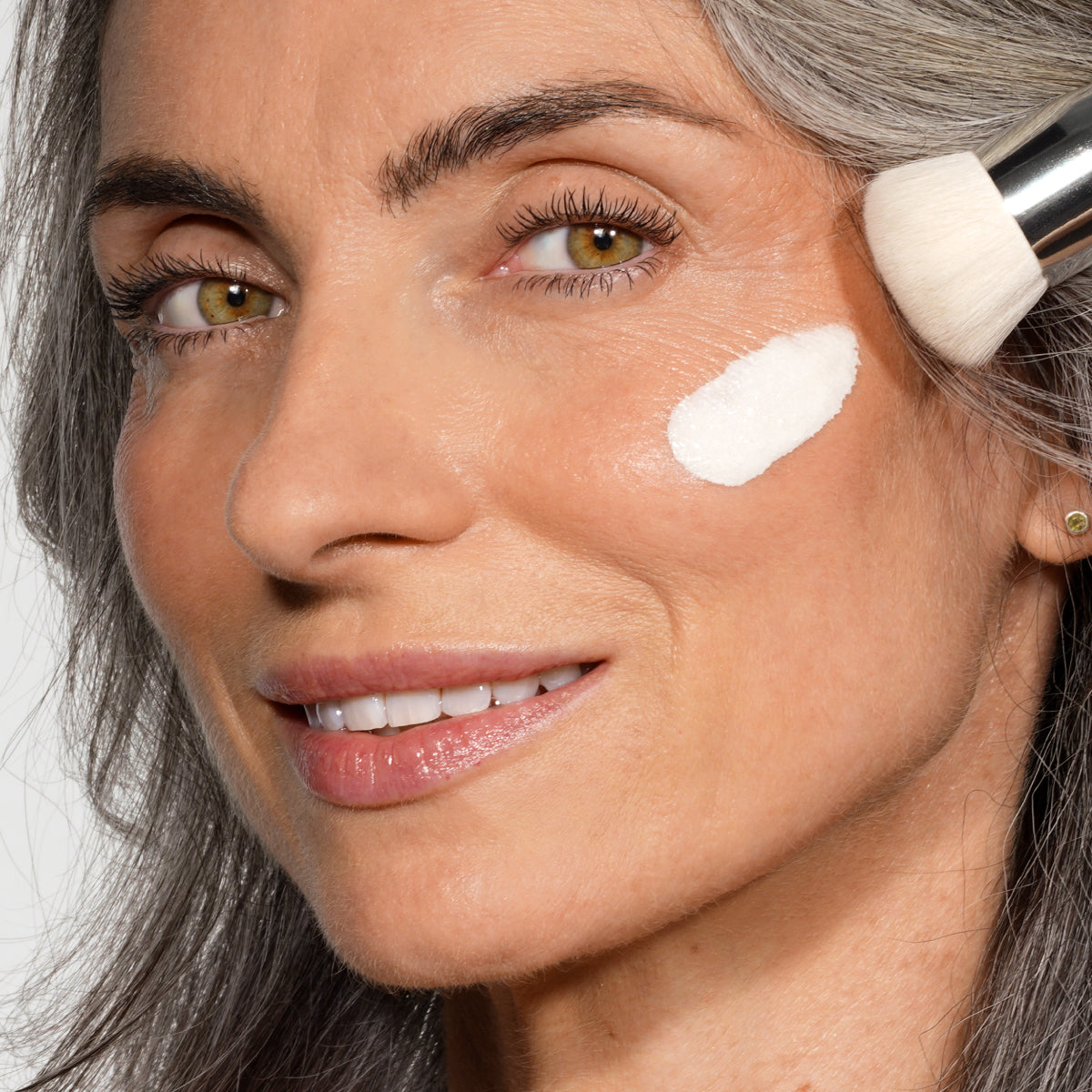 Eine Frau mit grauen Haaren lächelt, während sie mit einem Make-up-Pinsel weiße Creme auf ihre Wange aufträgt. Der Fokus des Bildes liegt auf ihrem Gesicht und dem Produkt, das sie gerade aufträgt.