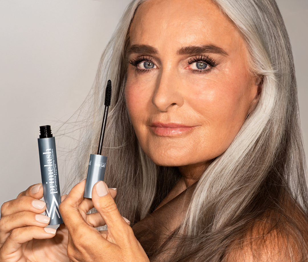 A woman with long gray hair and glowing skin holds an open mascara tube and wand near her face, showcasing her eye makeup and long eyelashes against a plain background.