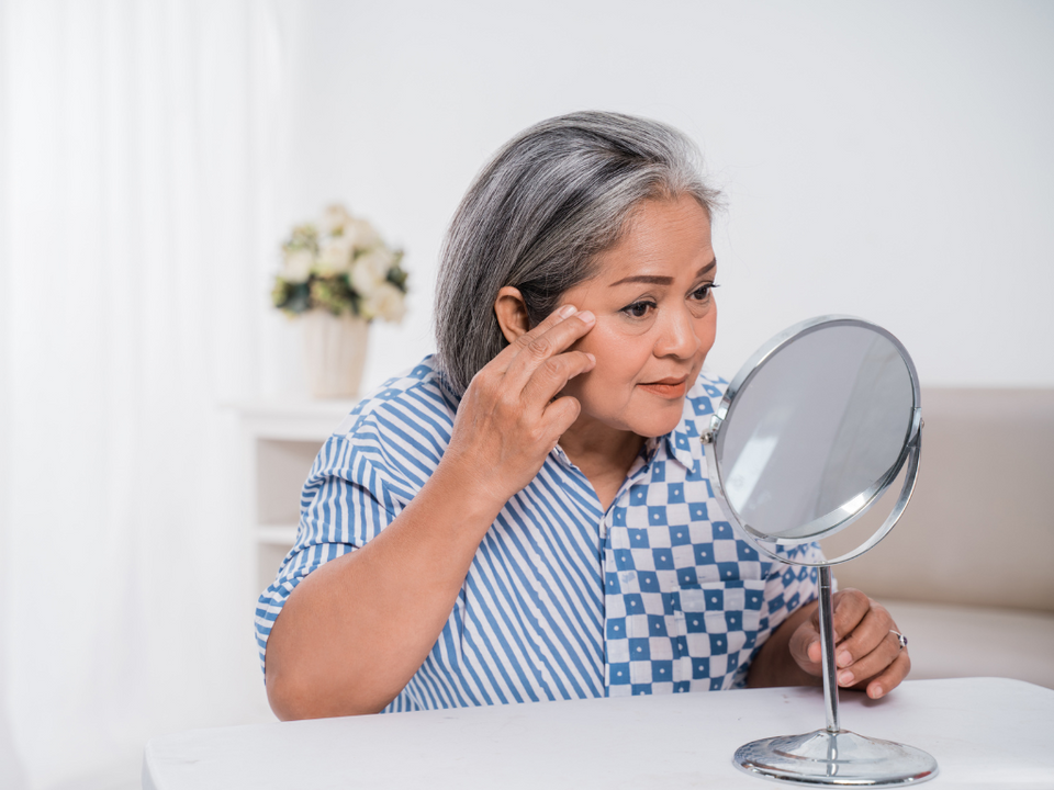 An older woman with gray hair examines her face in a round mirror, gently touching the skin near her eye. She is wearing a blue and white patterned shirt and sitting indoors at a white table.