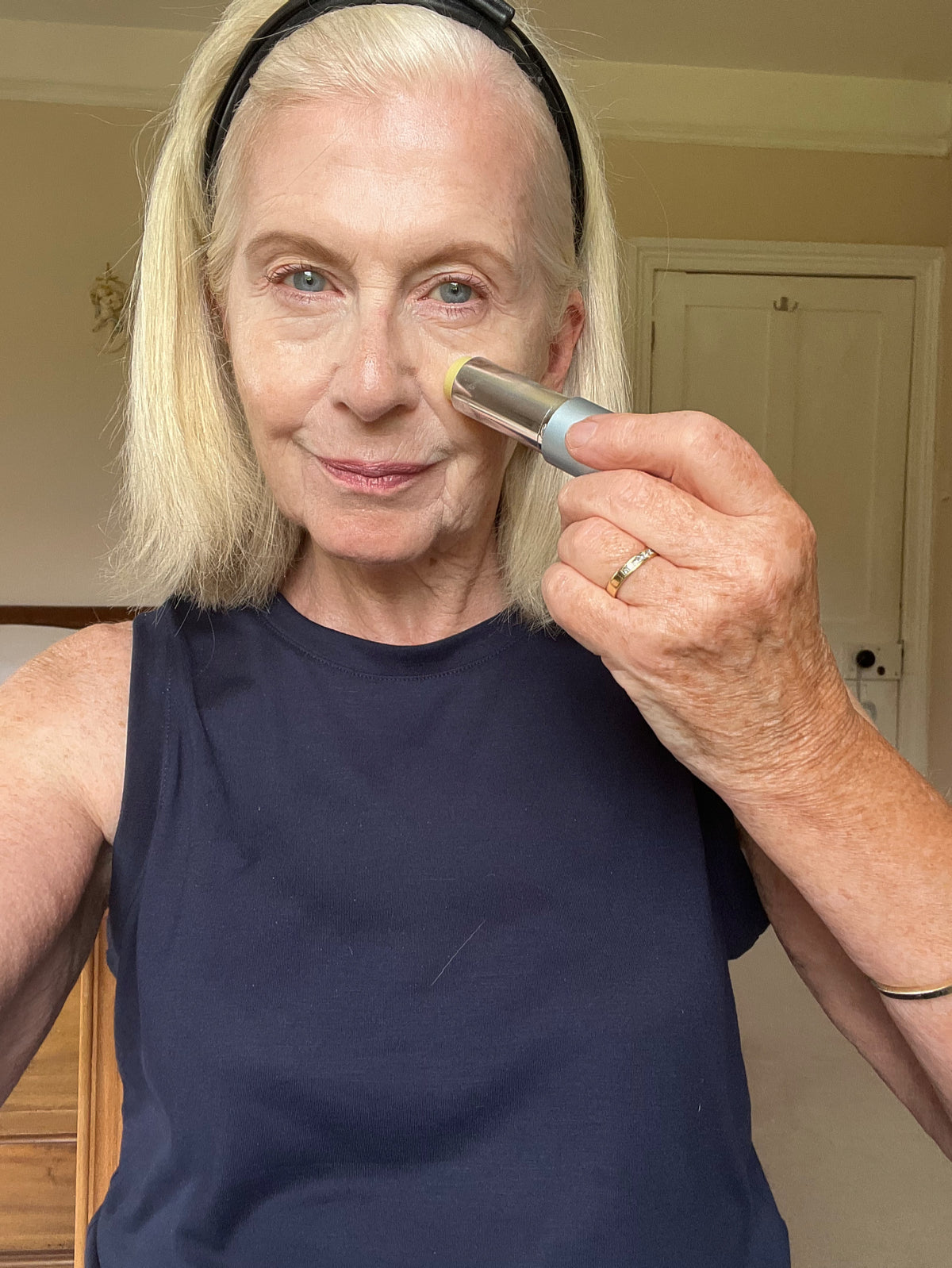 An older woman with blonde hair and a black headband is smiling and applying a makeup stick to her left cheek. She is wearing a sleeveless black top and a gold ring, and is standing indoors.