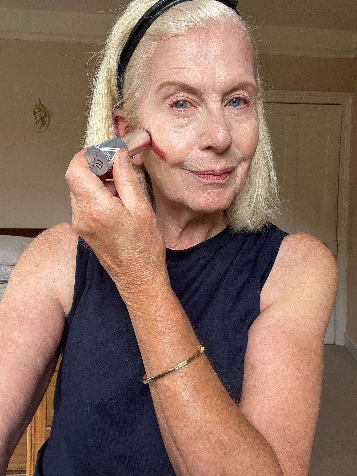 An older woman with short blonde hair applies makeup to her cheek using a stick blush. She is wearing a sleeveless black top, a headband, and a gold bracelet, and smiles softly while looking at the camera in a well-lit room.