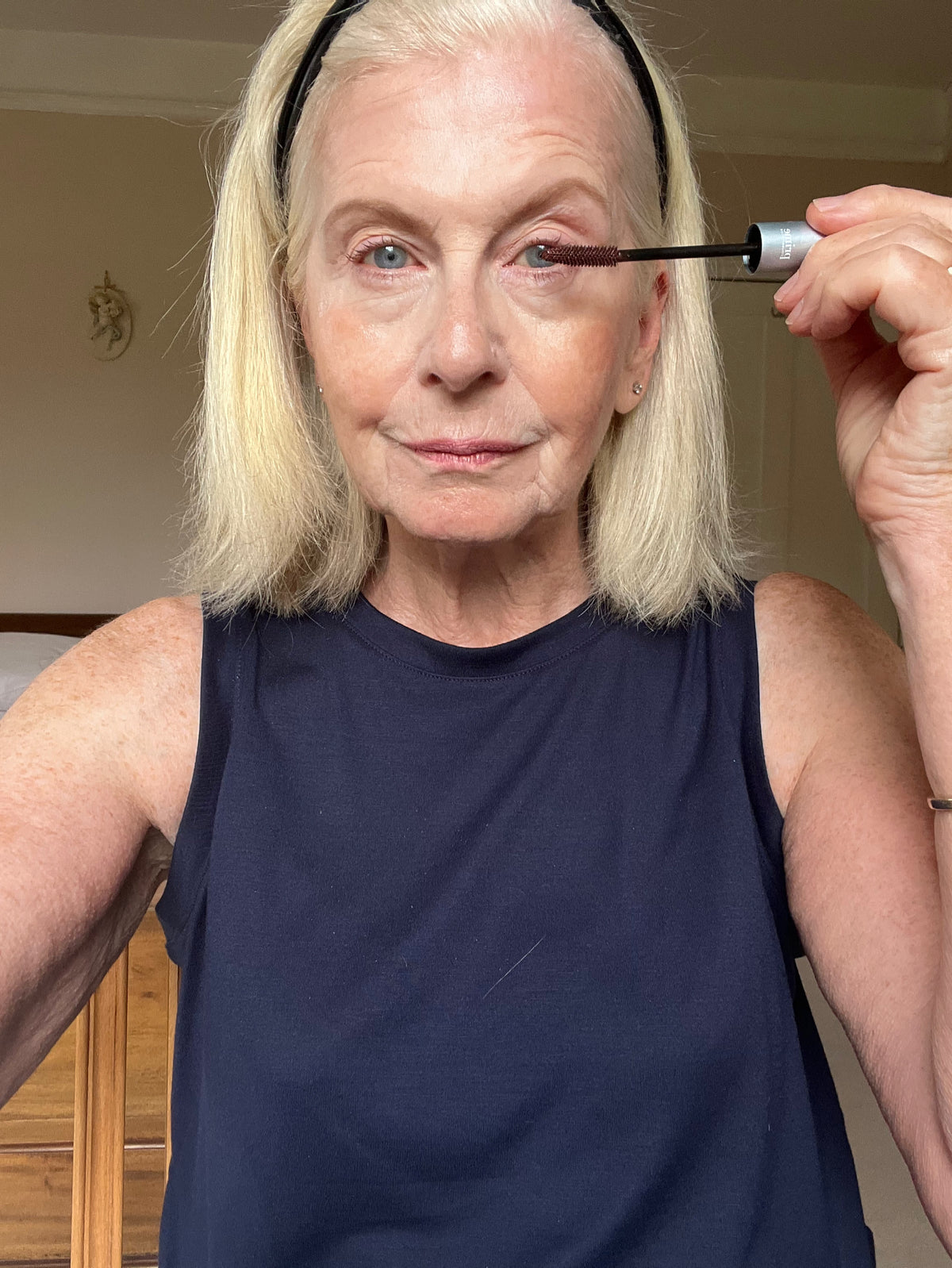 An older woman with shoulder-length blonde hair applies mascara to her upper eyelashes. She wears a sleeveless navy top and a black headband, looking directly at the camera in a well-lit room.