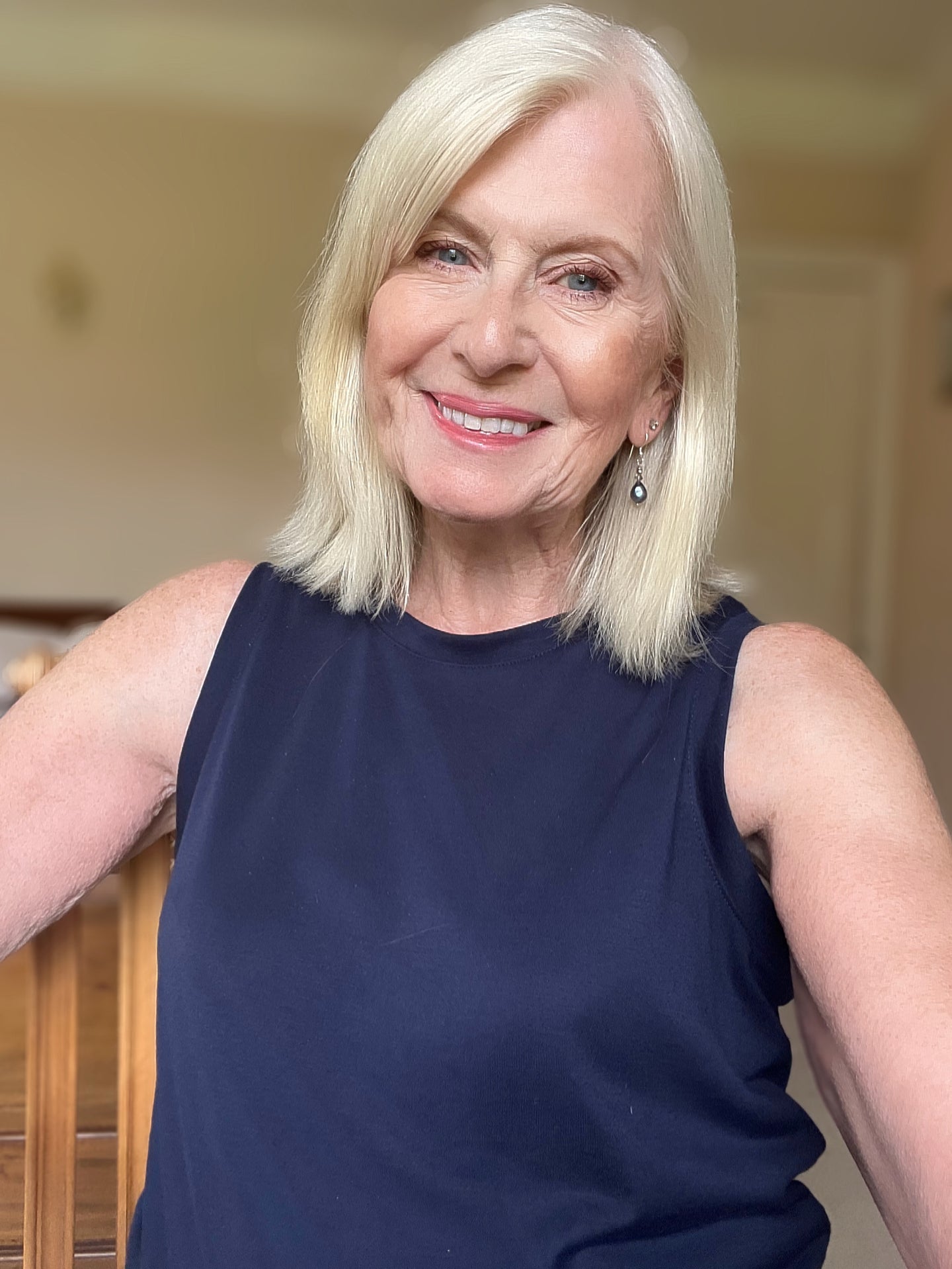 An older woman with shoulder-length blonde hair, wearing a sleeveless navy top and drop earrings, smiles warmly at the camera in a softly lit indoor setting.