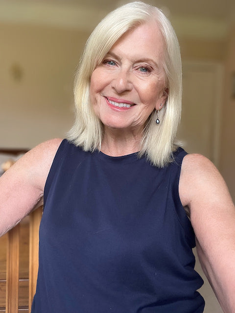 Smiling older woman with straight, shoulder-length blonde hair wearing a sleeveless navy top, standing indoors in a softly lit room.