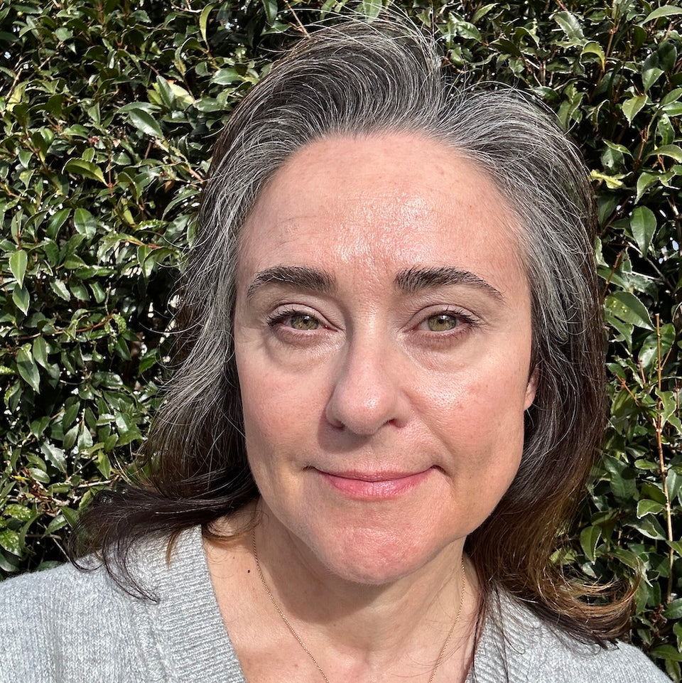 A woman with gray-brown hair and light skin stands outdoors in front of green leafy bushes, wearing a light gray top and looking at the camera with a gentle expression.