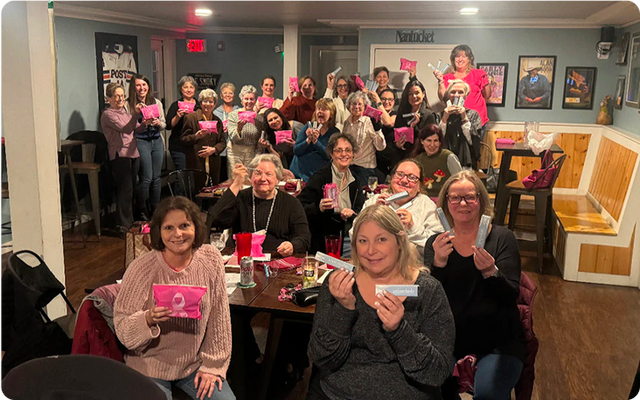 A group of women gathered in a restaurant, many smiling and holding up pink gift bags and tissue packs, posing together for a group photo around tables with drinks and snacks.