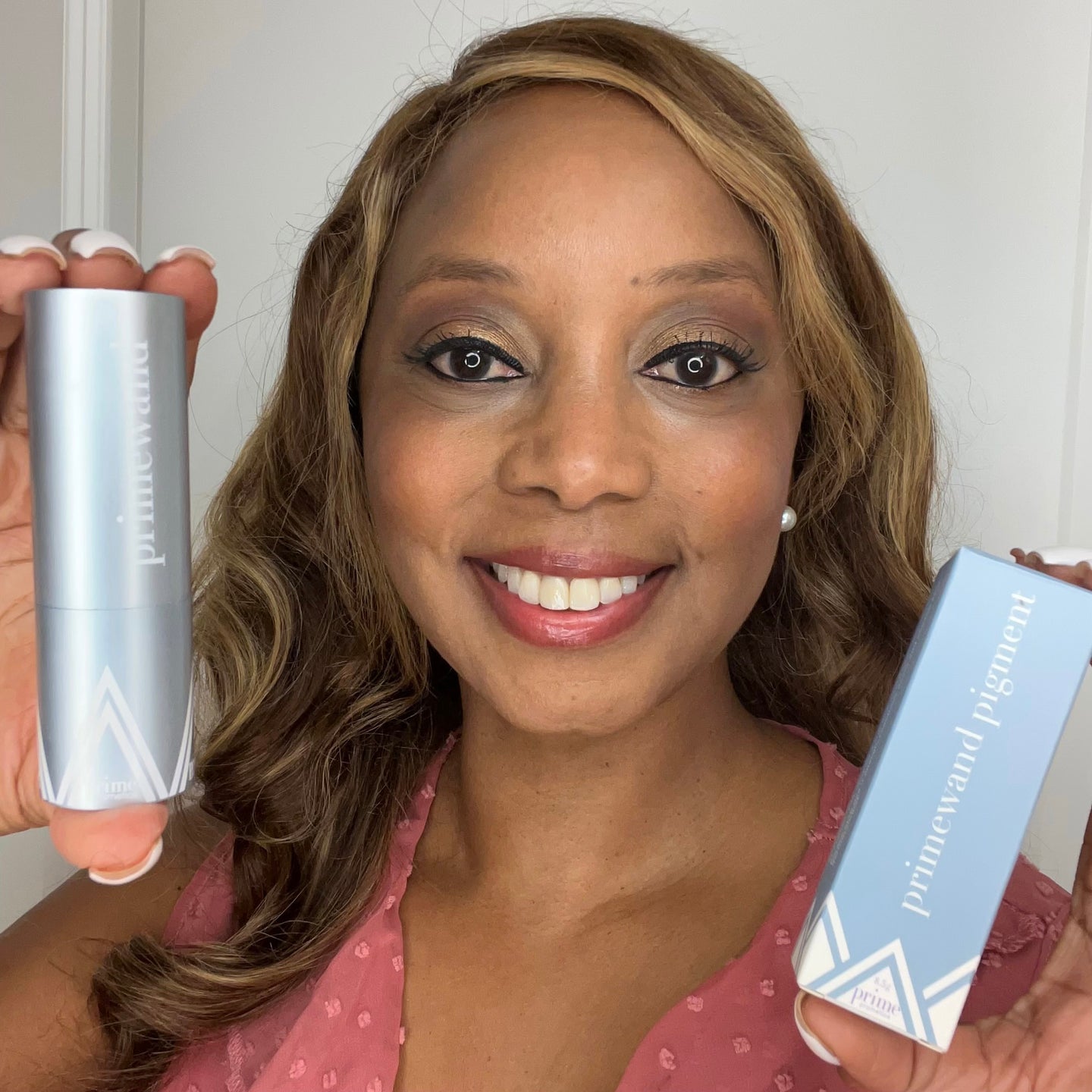 A smiling woman with long wavy hair holds up two silver-blue makeup products labeled primewand and primewand pigment in front of a white background. She wears a pink top and has natural makeup.
