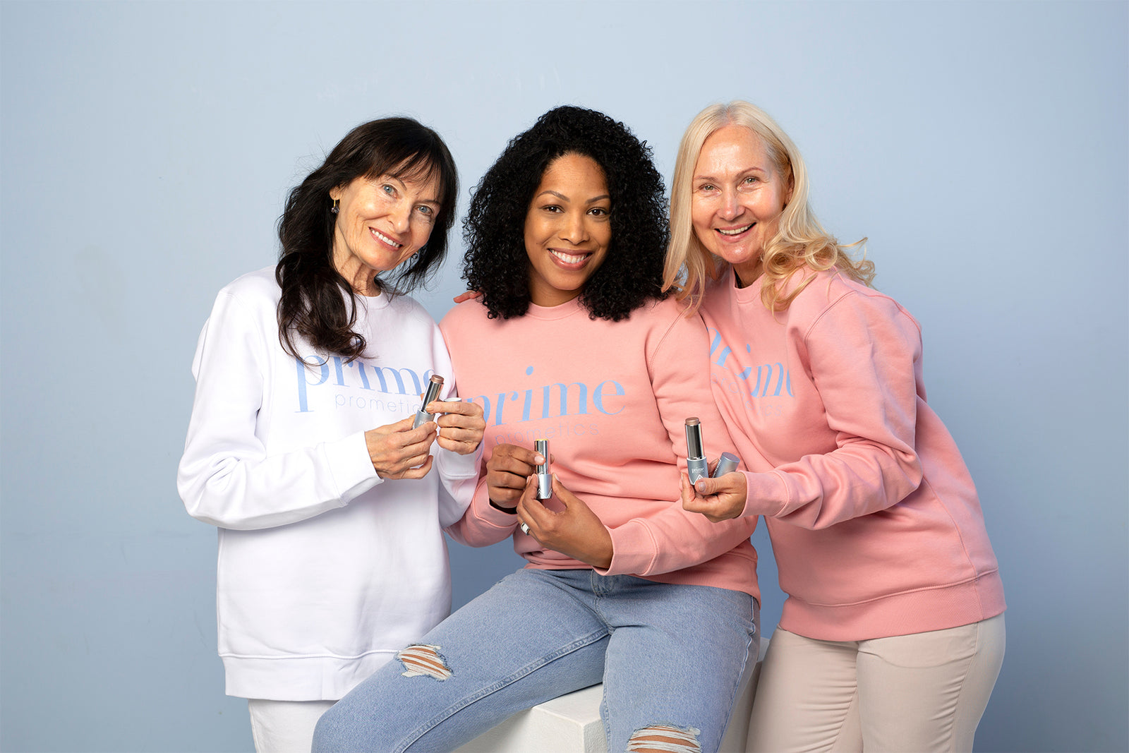 Three smiling women of different ages, wearing matching sweatshirts and holding skincare products, pose together against a light blue background. They appear friendly and confident, showcasing the items to the camera.