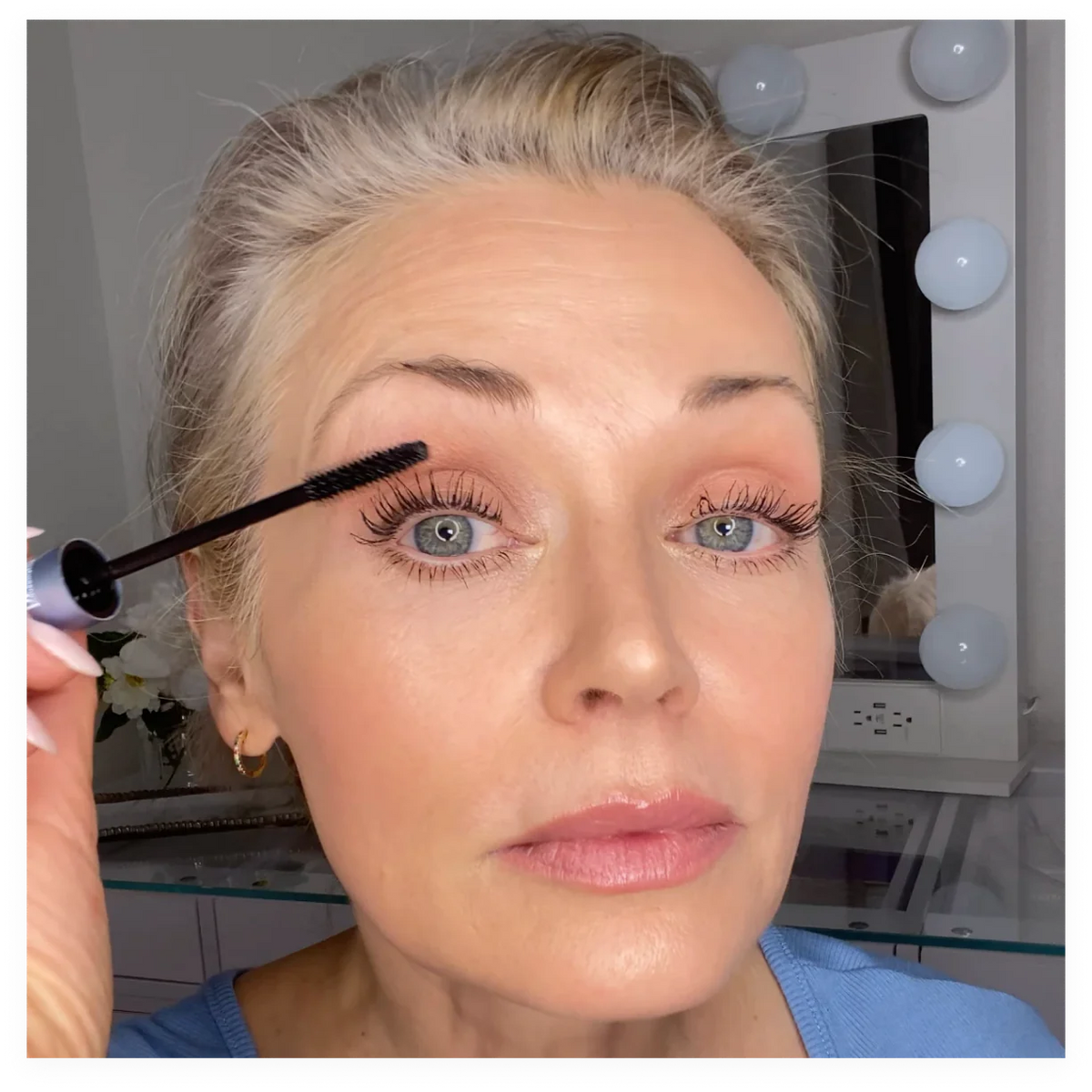 A woman with light hair and blue eyes applies mascara to her upper eyelashes. She is wearing a blue top and is in a well-lit room with a mirror surrounded by light bulbs in the background.