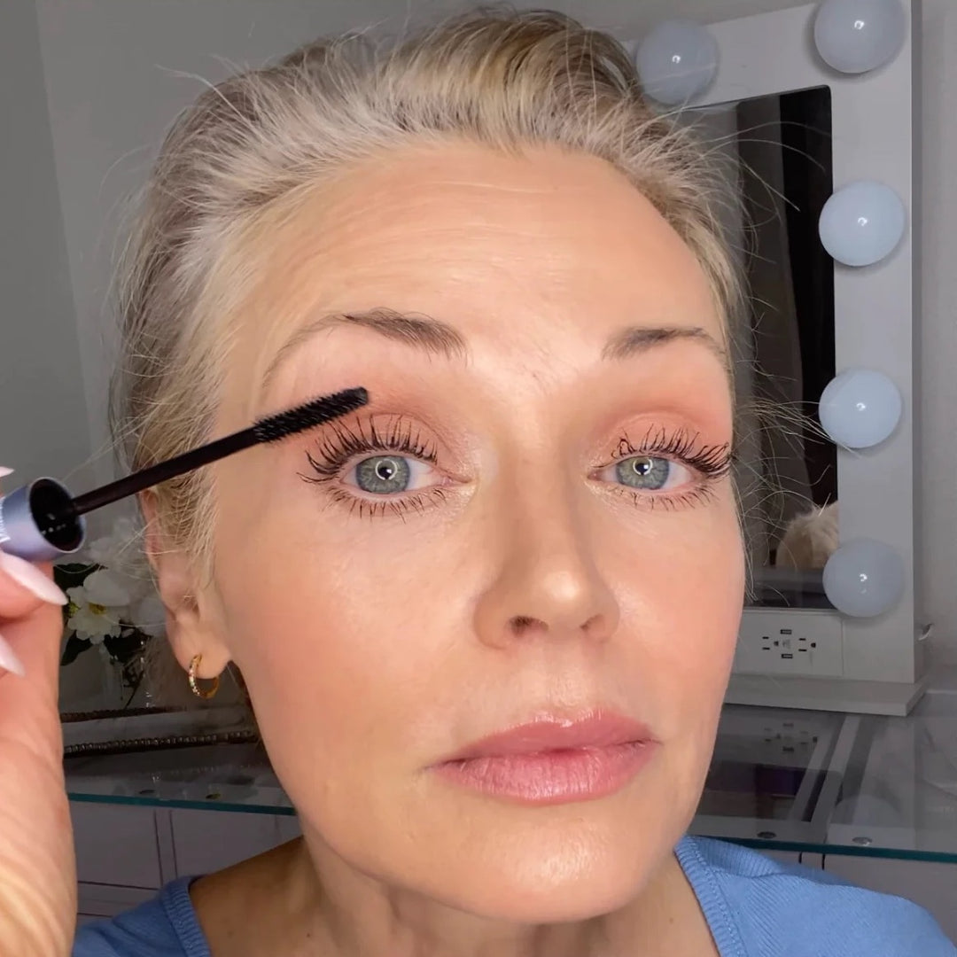 A woman with light hair applies mascara to her eyelashes in front of a mirror with round lights. She is wearing a blue top and gold hoop earrings, and looks directly at the camera.