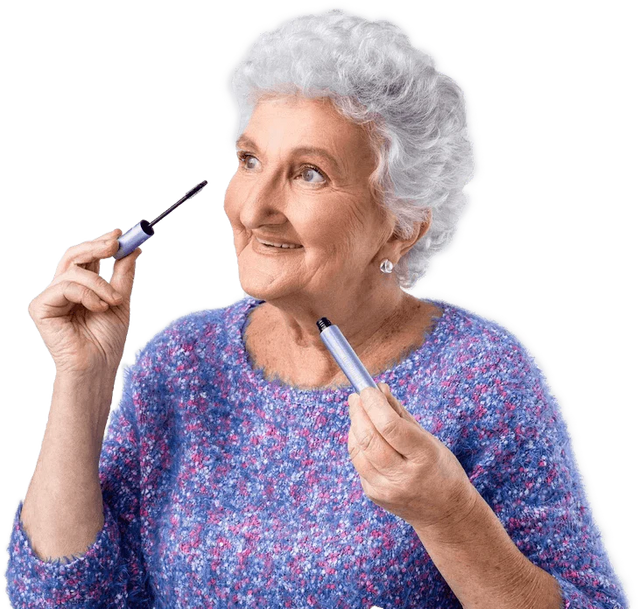 An elderly woman with curly white hair and a purple sweater is smiling while holding a mascara wand near her eye, appearing to apply makeup.