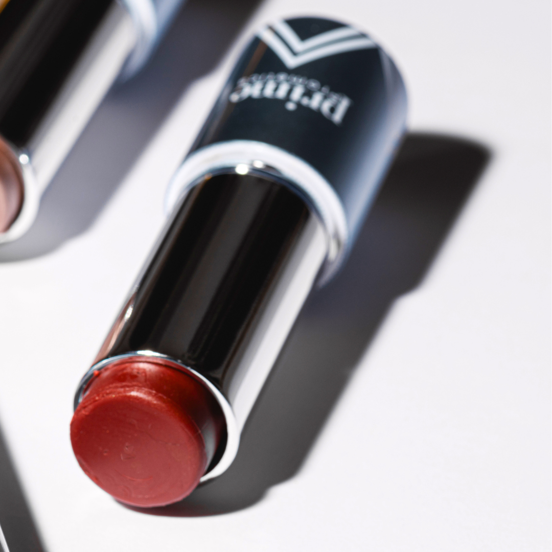 A close-up of an open lipstick with a reddish shade, lying on a white surface. The lipstick tube is black and silver with a hint of branding visible on the cap.
