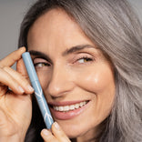 A smiling woman with long gray hair holds a blue tube of PrimePrometics™ 2pcs PrimeEyes Eyeshadow Matte near her face, showing off her clear skin and natural makeup.