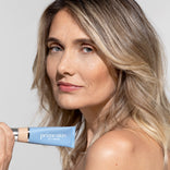 A woman with blonde hair holds a blue tube of Primeskin CC+ Cream near her face, looking confidently at the camera against a plain background.