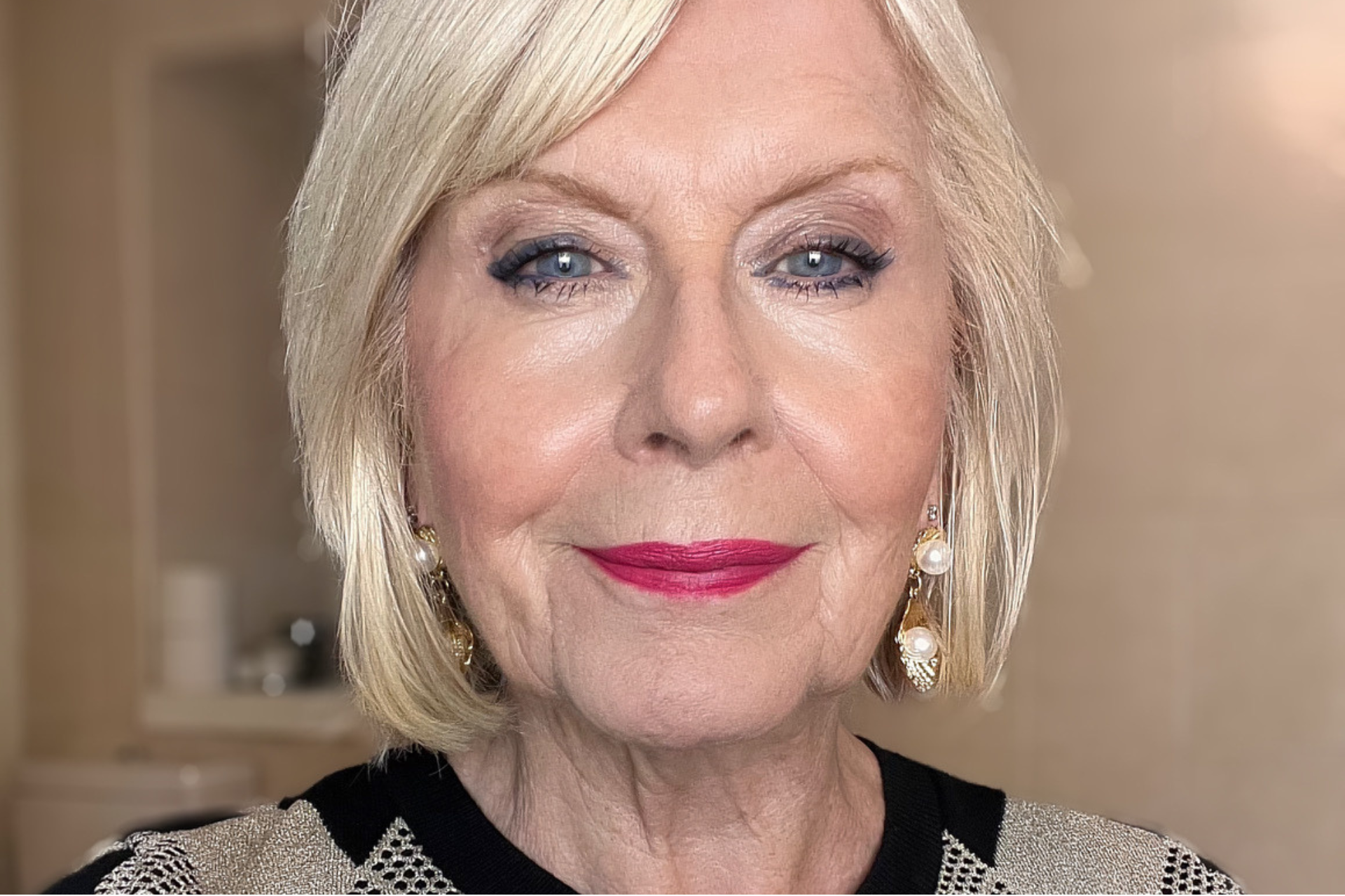 A close-up of an older woman with short, blonde hair, blue eyes, and pink lipstick. She is wearing gold and pearl drop earrings and a patterned top. The background is softly blurred.