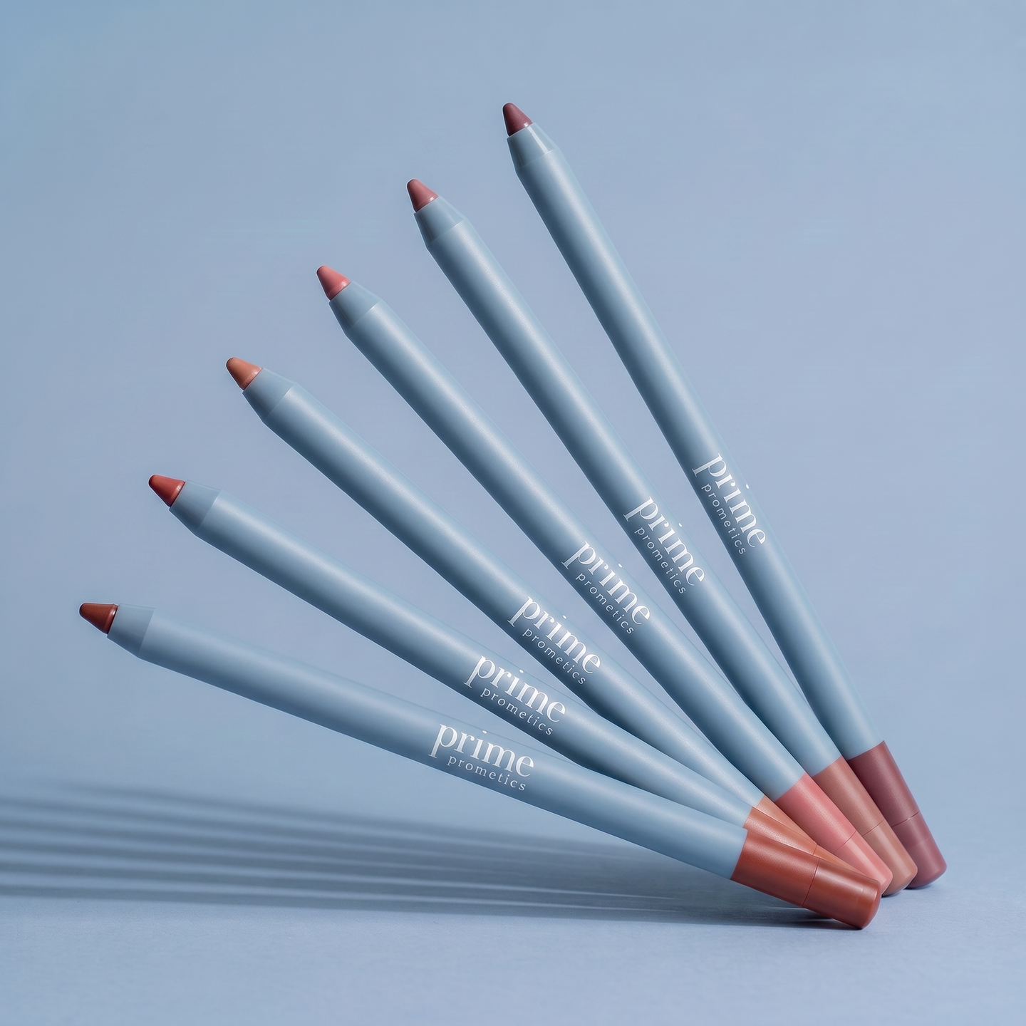 Five blue makeup pencils labeled prime paramedics are arranged in a fan shape on a blue surface, with each pencil showing a different shade of lip color at the tip.